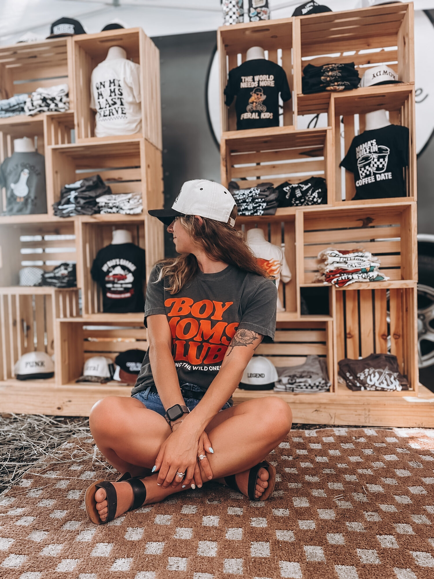 Young woman sitting cross-legged on a patterned rug in a clothing store, surrounded by wooden shelves filled with folded clothes.