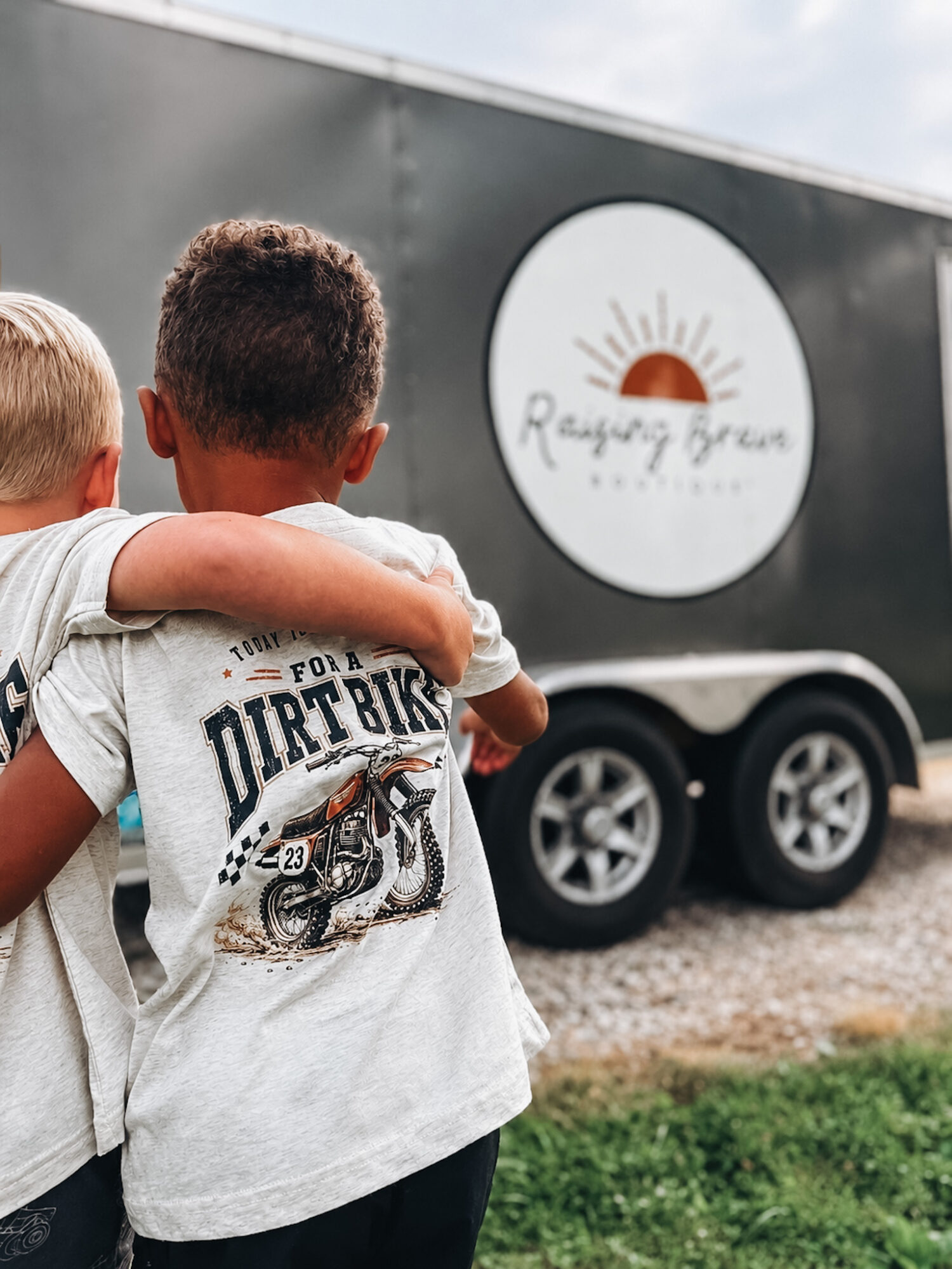 Two children hugging outdoors near a trailer with a circular logo featuring a sun and text.