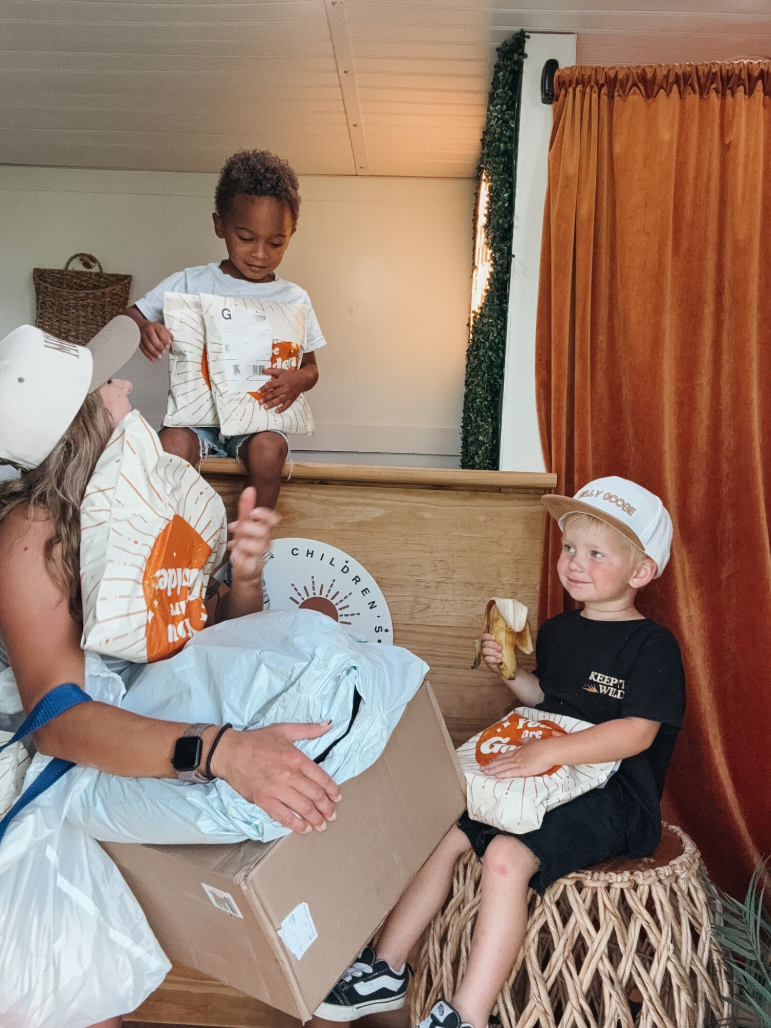 Two children, one standing on a bed and one sitting on a stool, sharing snacks in a room with curtains.
