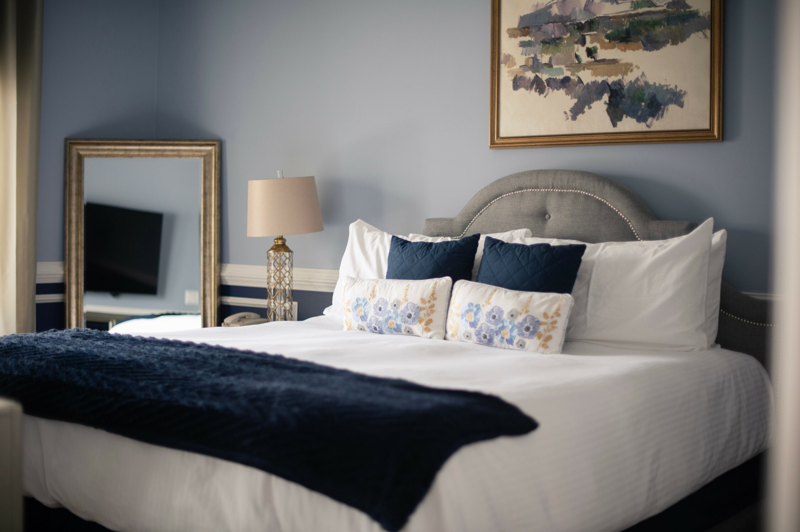 A neatly made bed with white and navy pillows, a navy blanket, a bedside lamp, a mirror, and artwork on a blue wall.