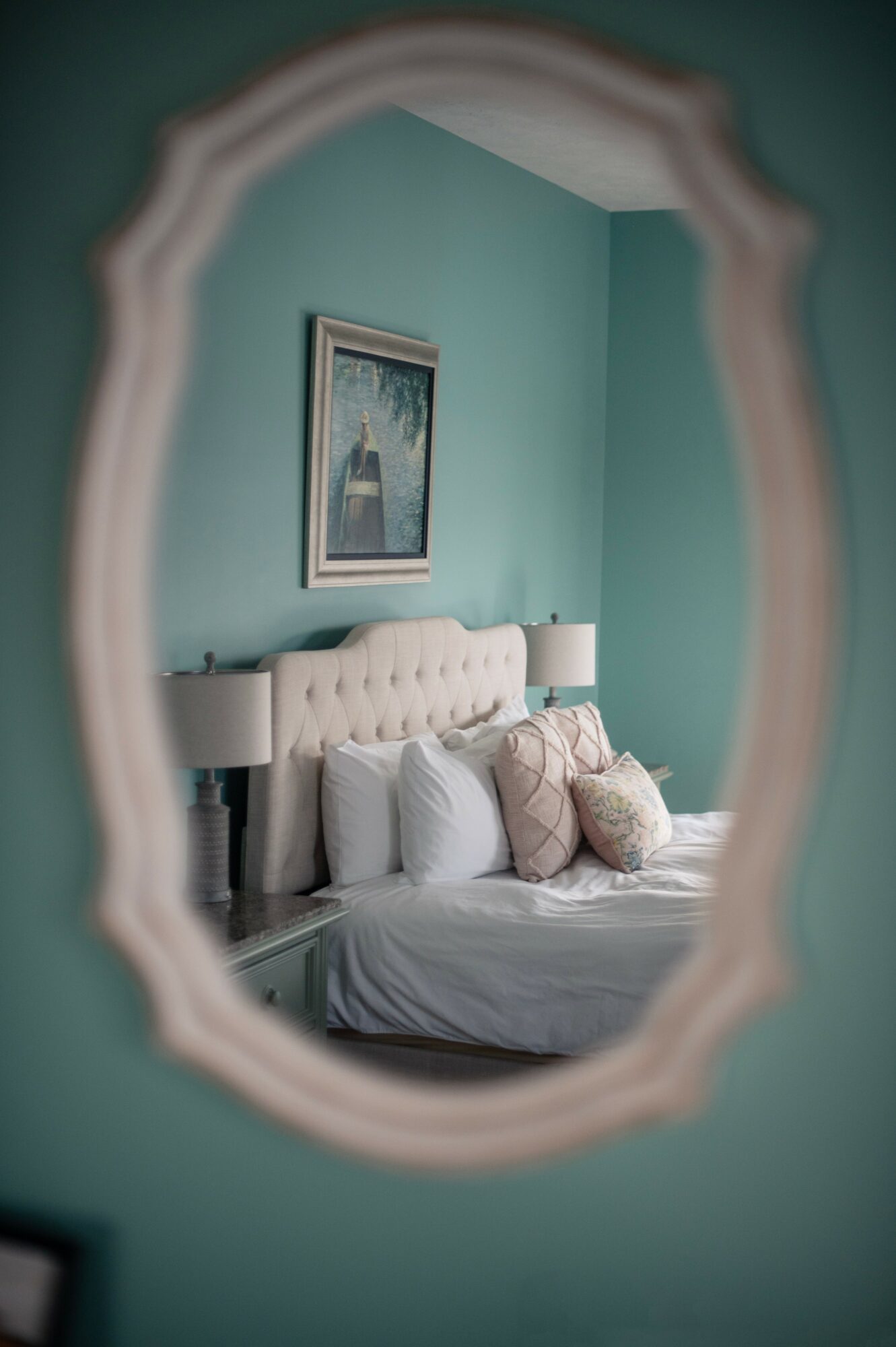 View of a bedroom through an ornate mirror, showing a bed with pillows, a painting on the wall, and teal walls.