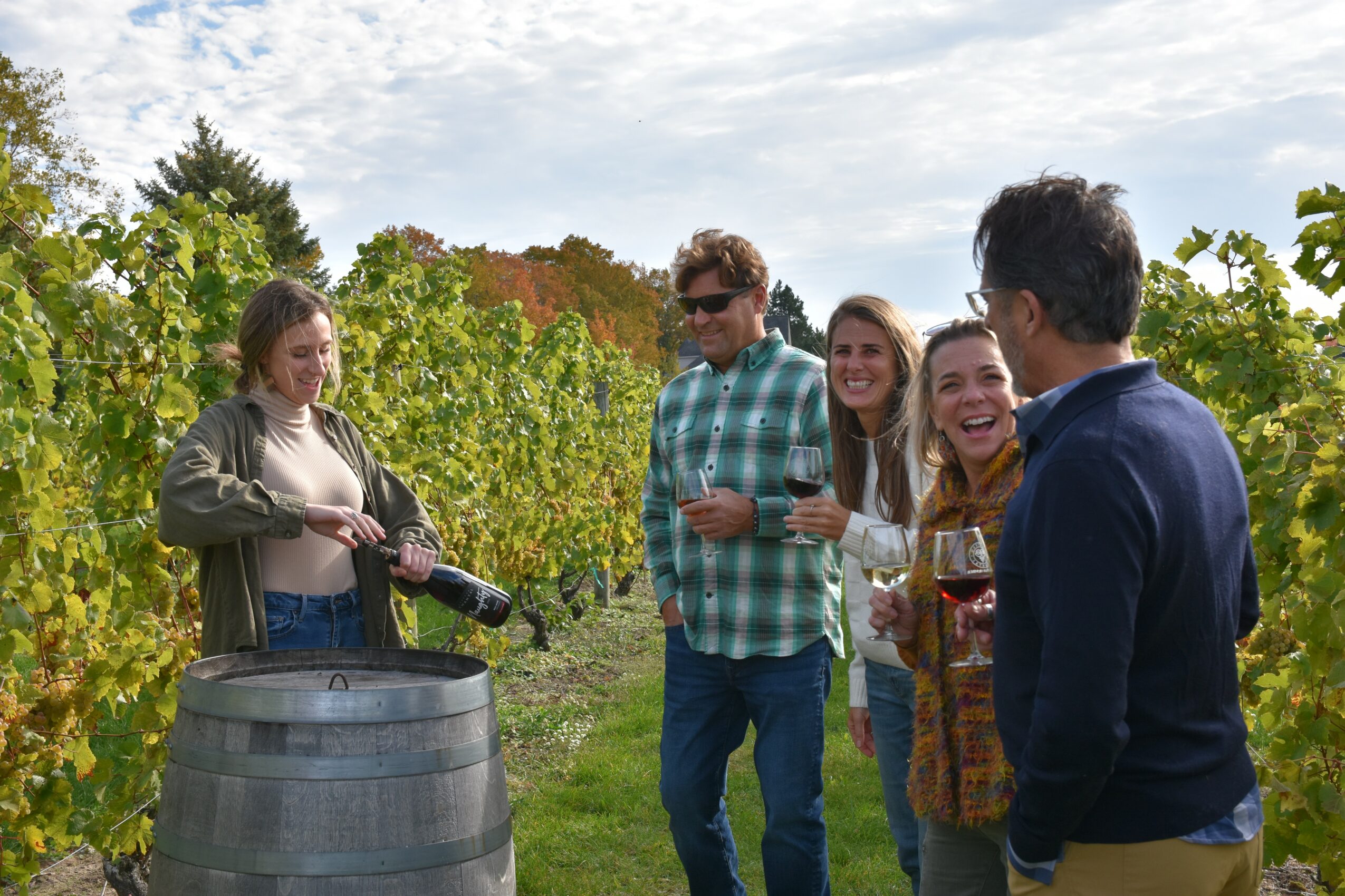 Group of five people outdoors in a vineyard, smiling and talking, with a woman pouring wine from a bottle into glasses.