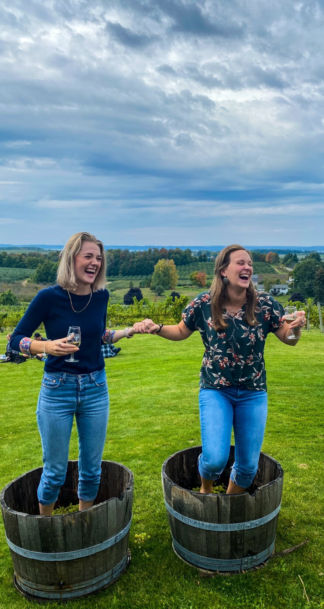 Two women standing in wooden barrels outdoors, holding hands and laughing, with a grassy field and cloudy sky in background.