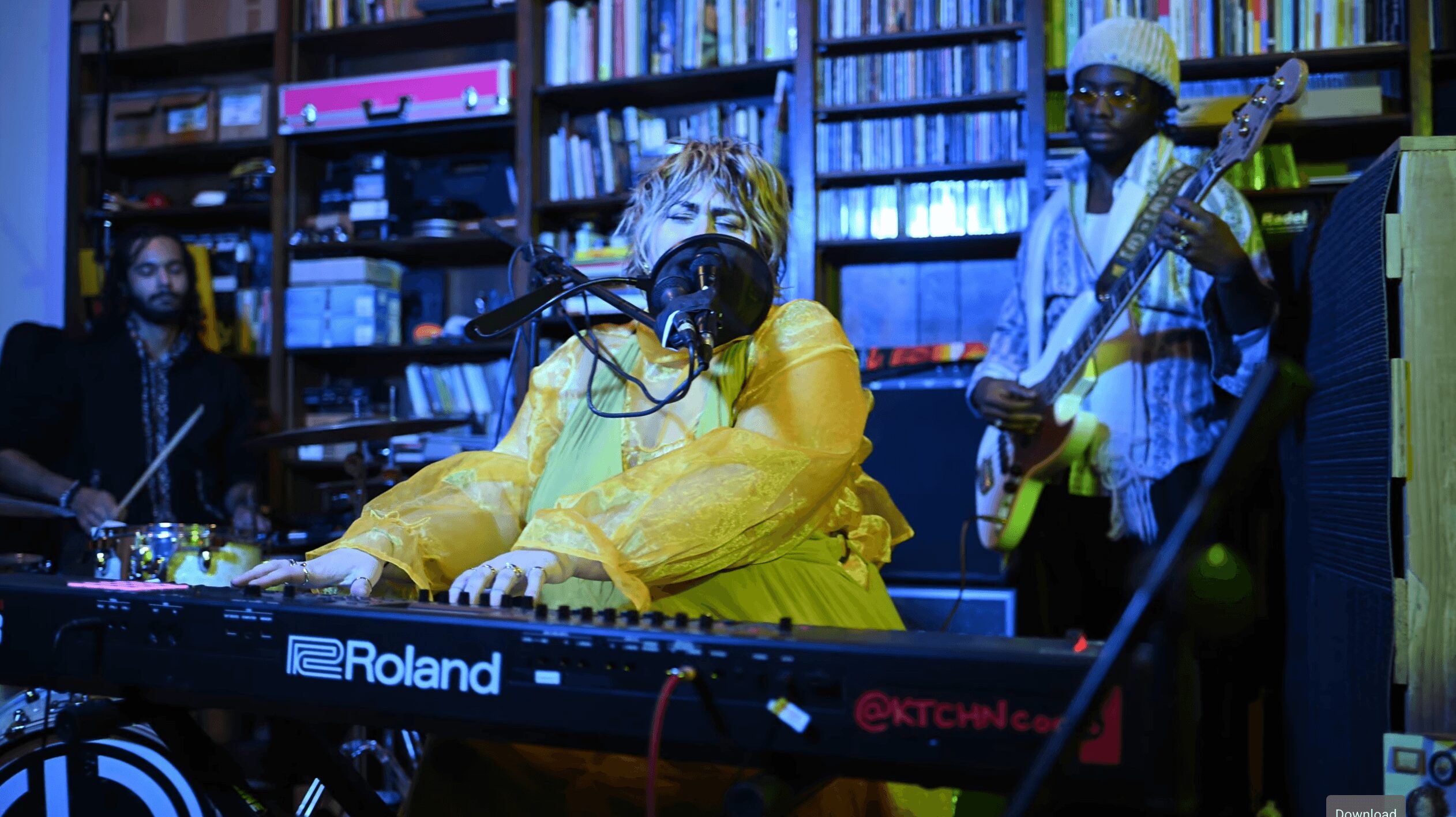 Musicians perform in a room with bookshelves, a woman plays keyboard, and two men play guitar.