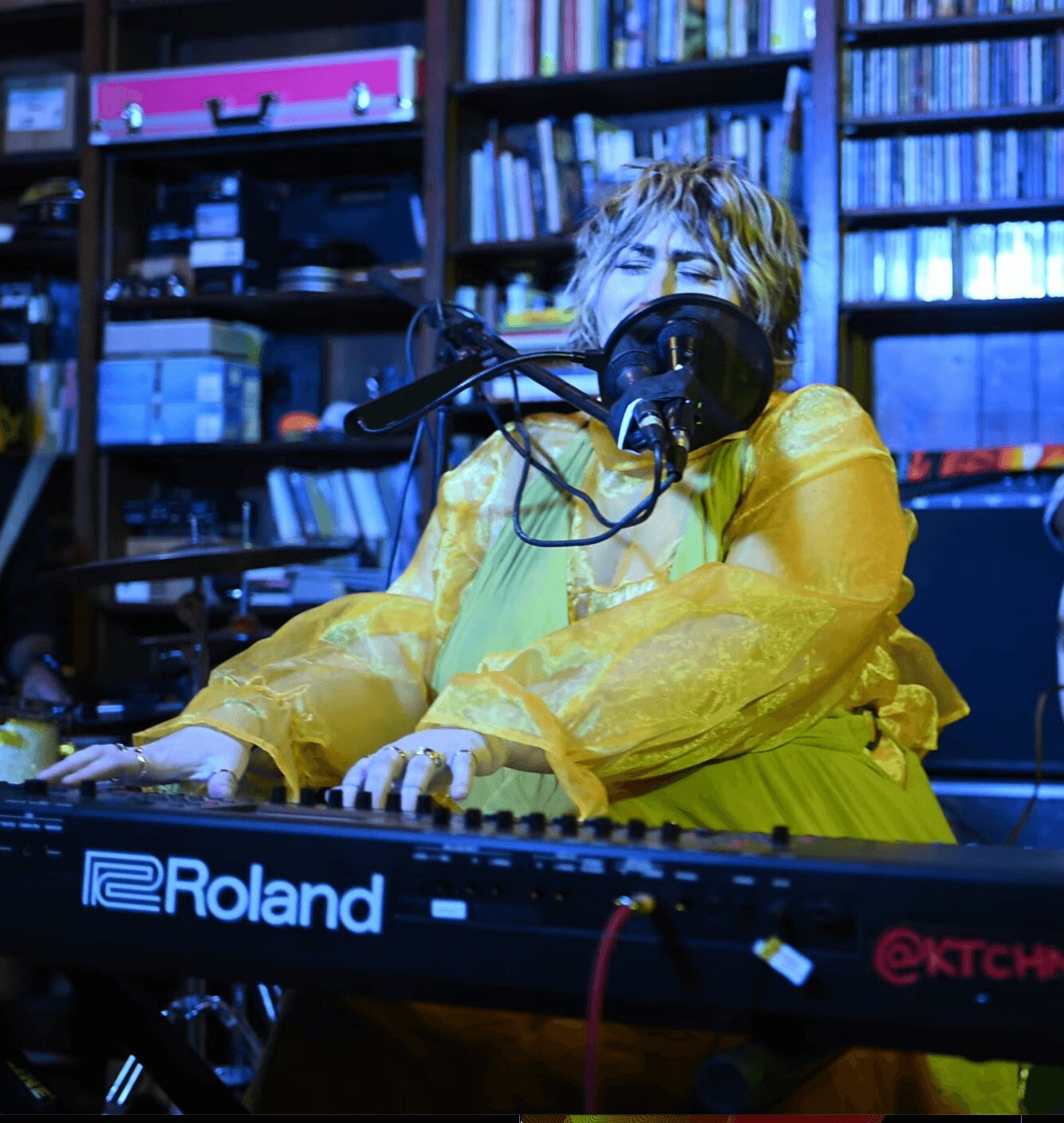 Person playing a Roland keyboard with a microphone and headphones in a room with shelves of books and supplies