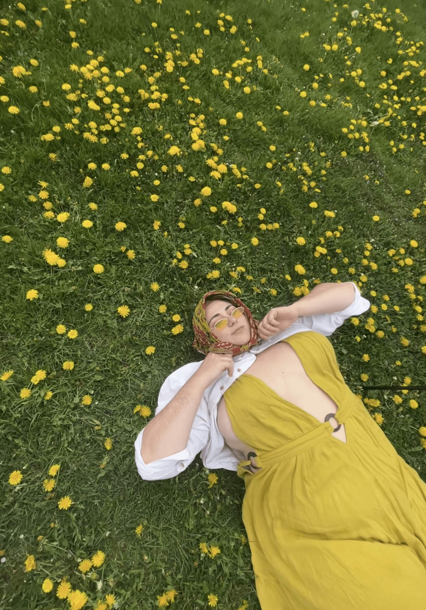 Woman lying on grass with yellow flowers, wearing a yellow dress and white top, looking up.