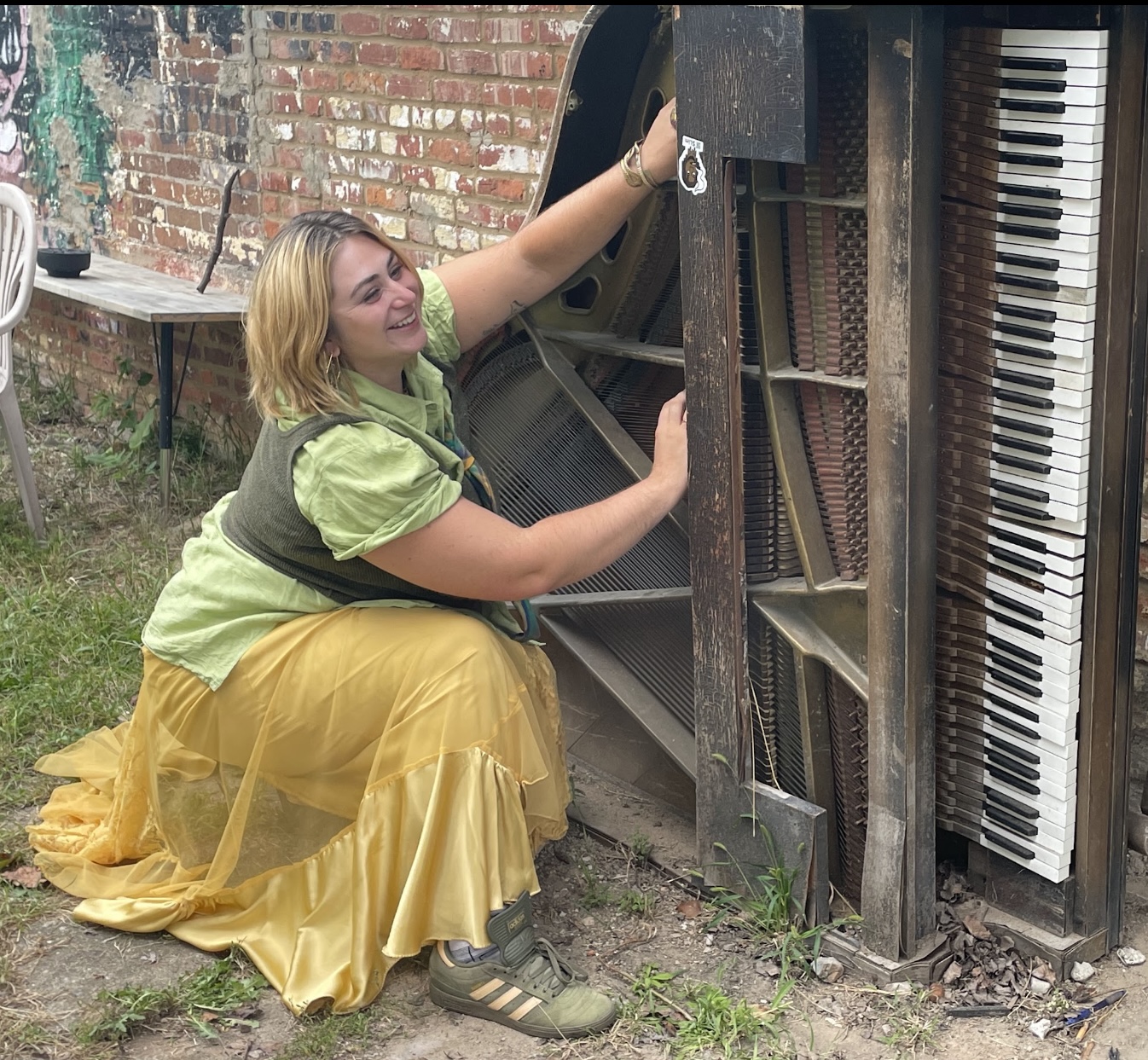 Person with blonde hair in a green top and yellow skirt interacts with an old upright piano outdoors.