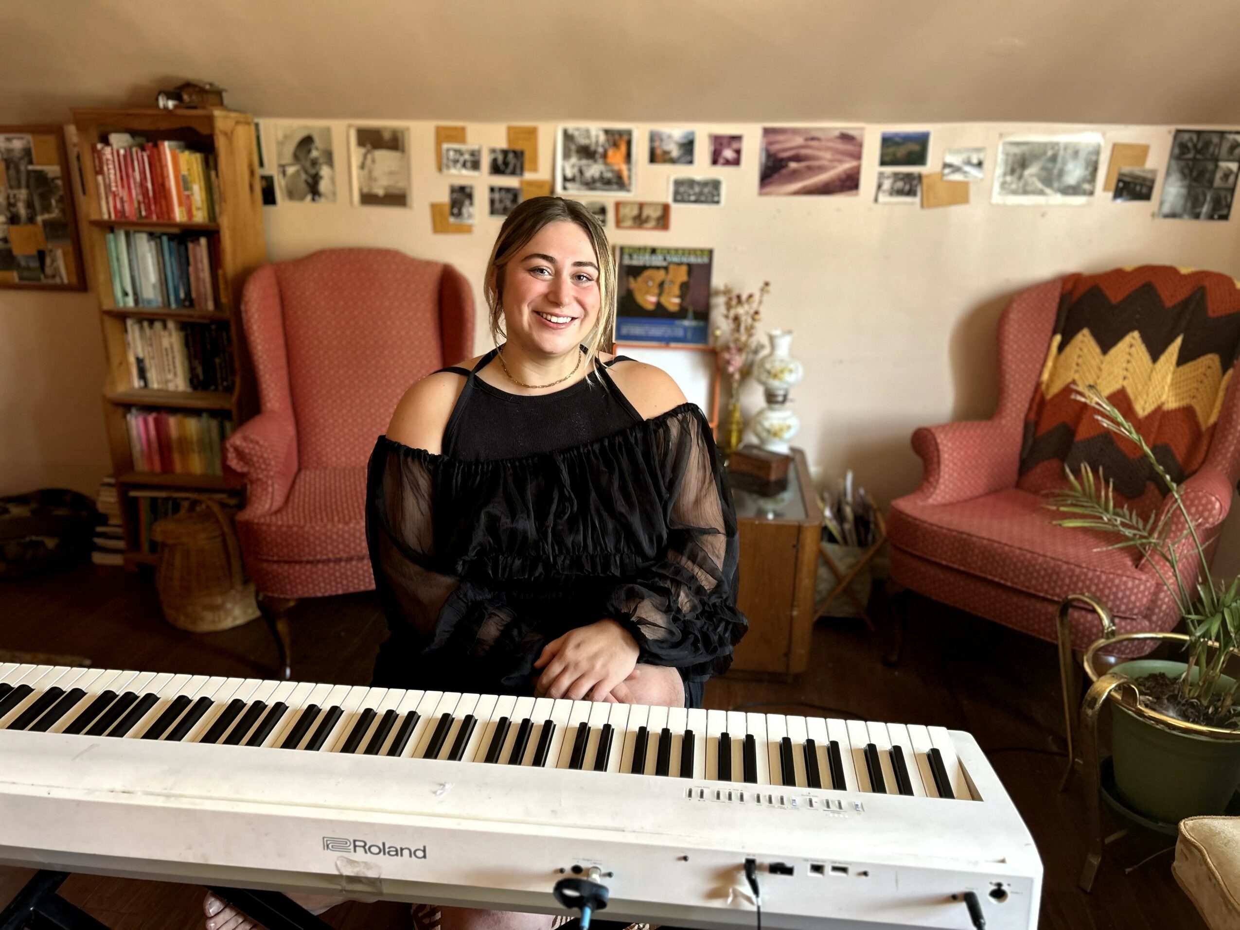 Young woman with shoulder-length hair smiling, sitting behind a keyboard in a cozy room with bookshelves and photos on the wall.