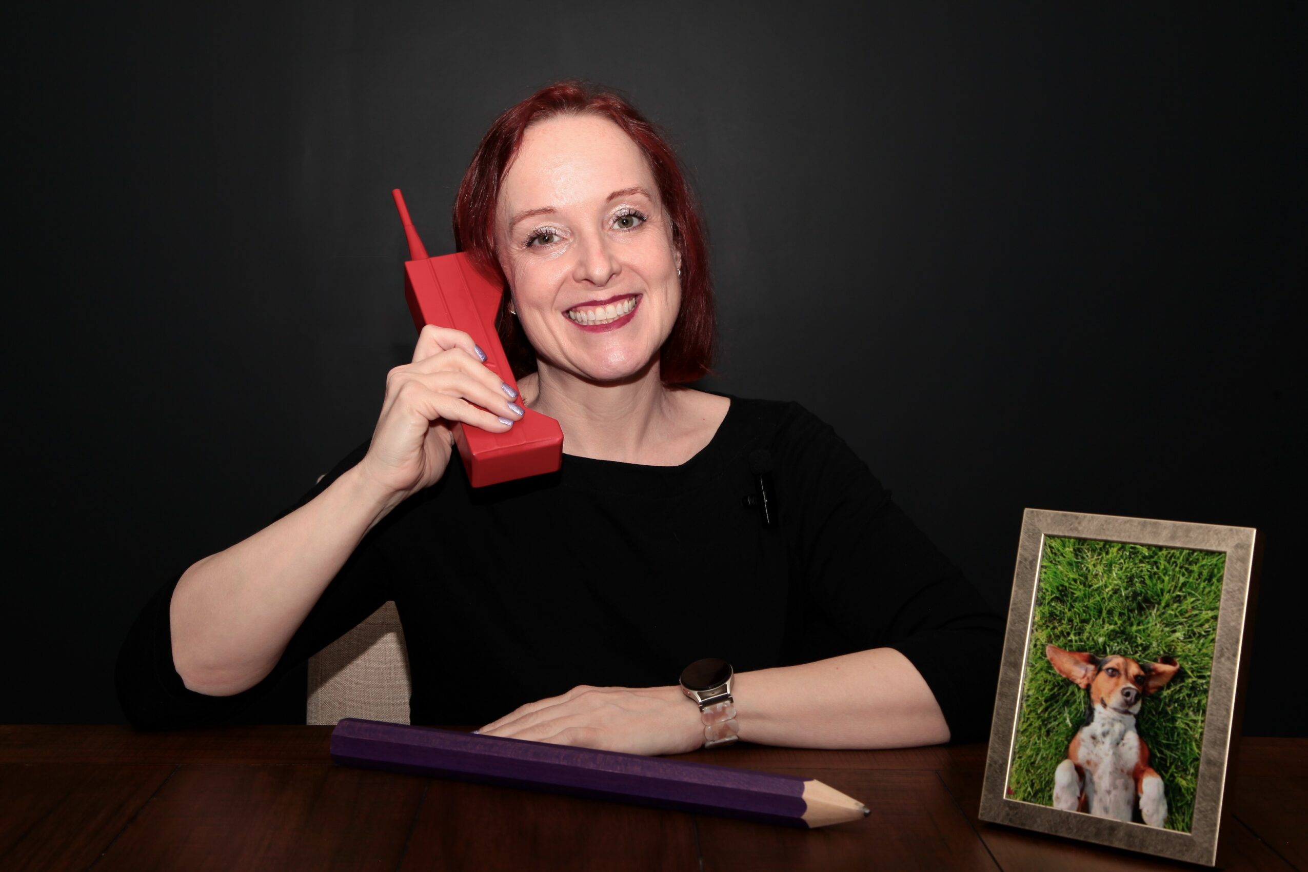 Woman with red hair smiling, holding a red phone to her ear, sitting at a table with a framed dog photo, black background