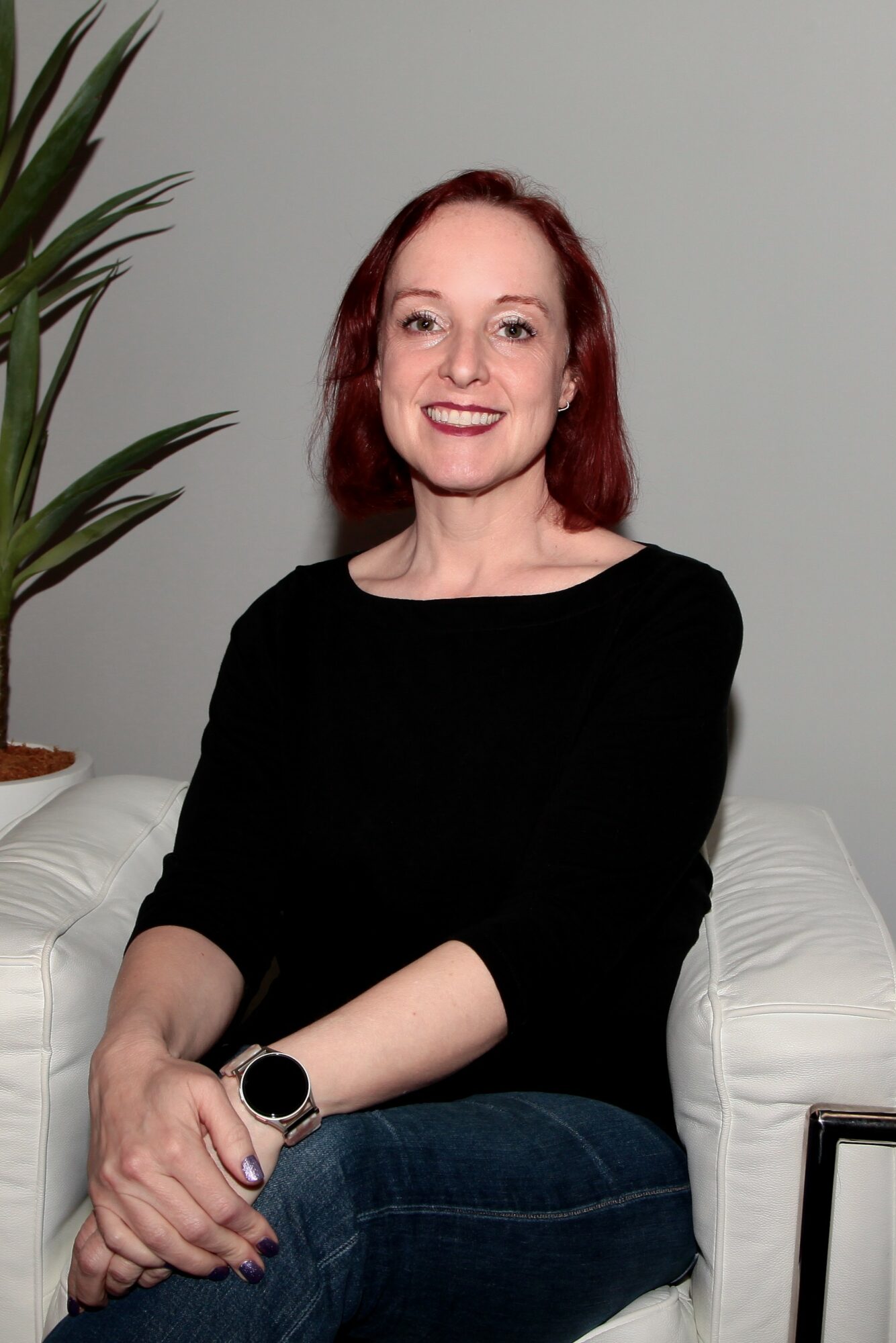 Woman with red hair smiling, wearing a black top, sitting on a white couch, with a plant nearby.