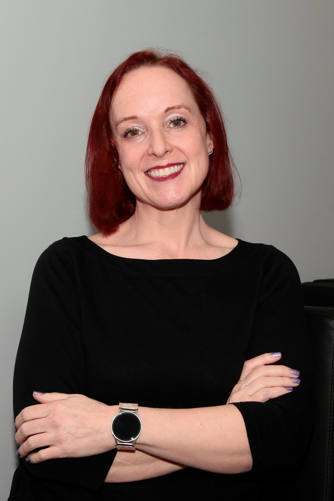 Woman with red hair smiling, arms crossed, wearing a black top and watch, against a plain background.