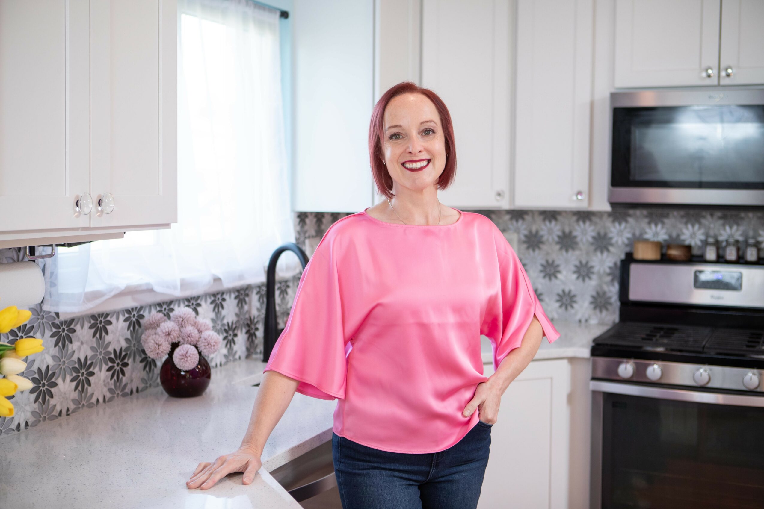 Woman with red hair smiling in a kitchen, wearing a pink top and dark pants, standing near a countertop.