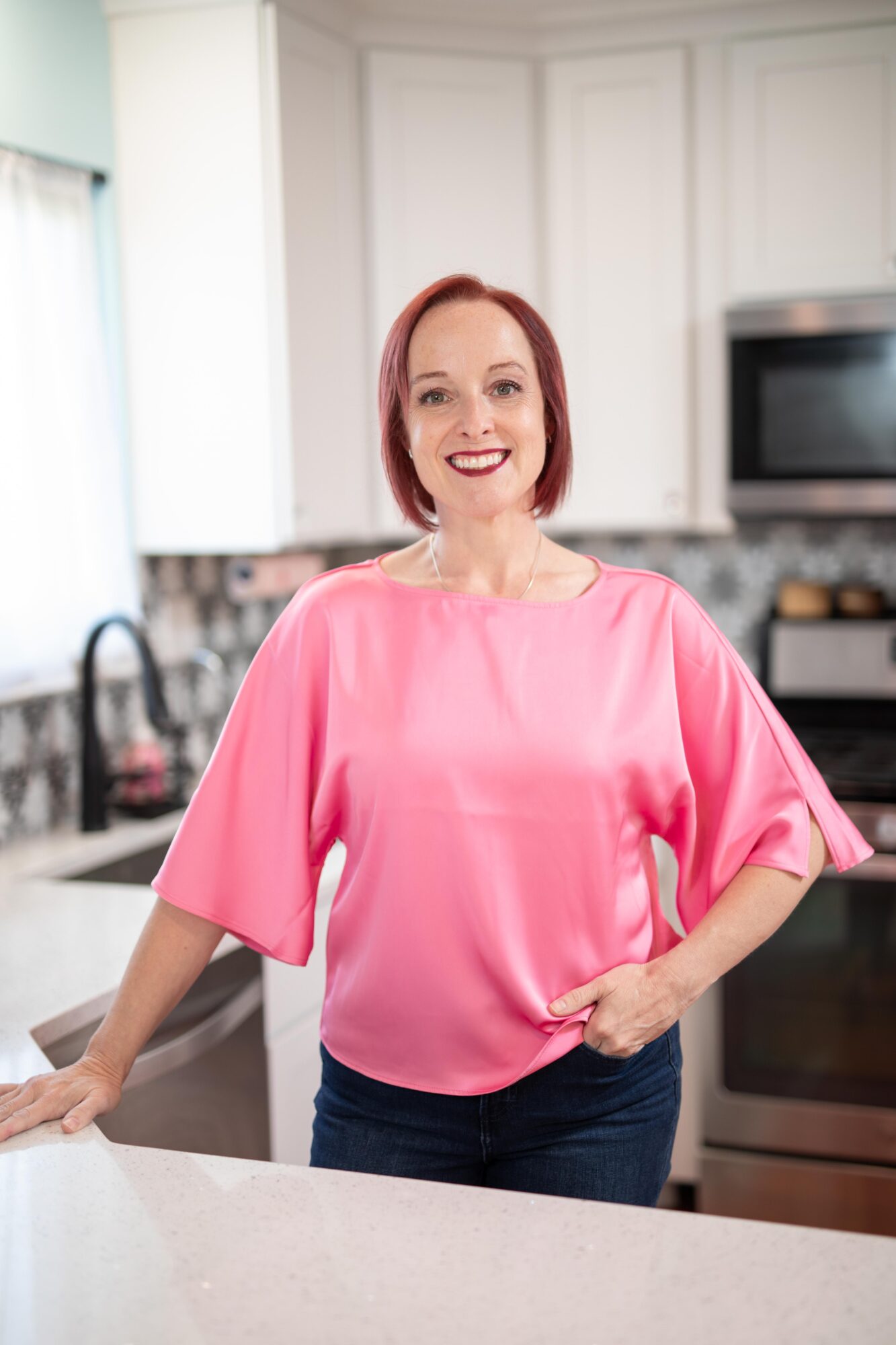 Woman with red hair smiling in a kitchen, wearing a pink blouse and dark pants.