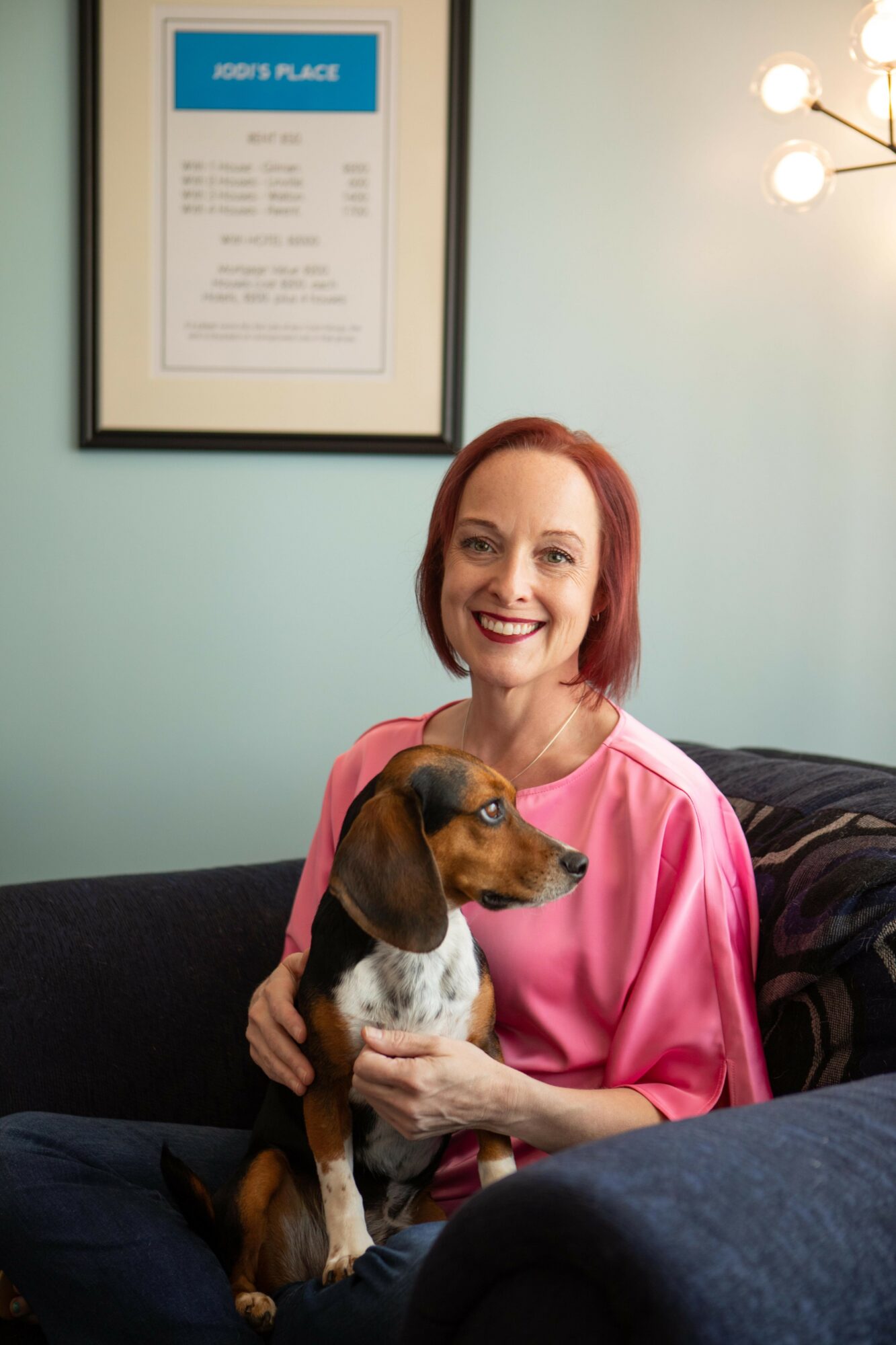 Woman with red hair smiling, holding a small brown and white dog, sitting on a dark sofa in a room with light green walls.