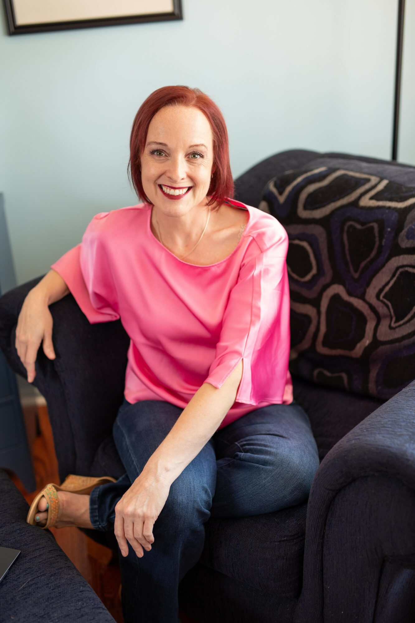 Smiling woman with red hair sitting on a dark sofa, wearing a pink top and jeans, in a room with light-colored walls.