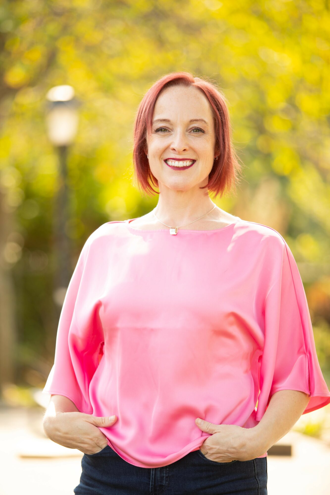 Smiling woman with short pink hair wearing a pink blouse outdoors with yellow-green foliage background.
