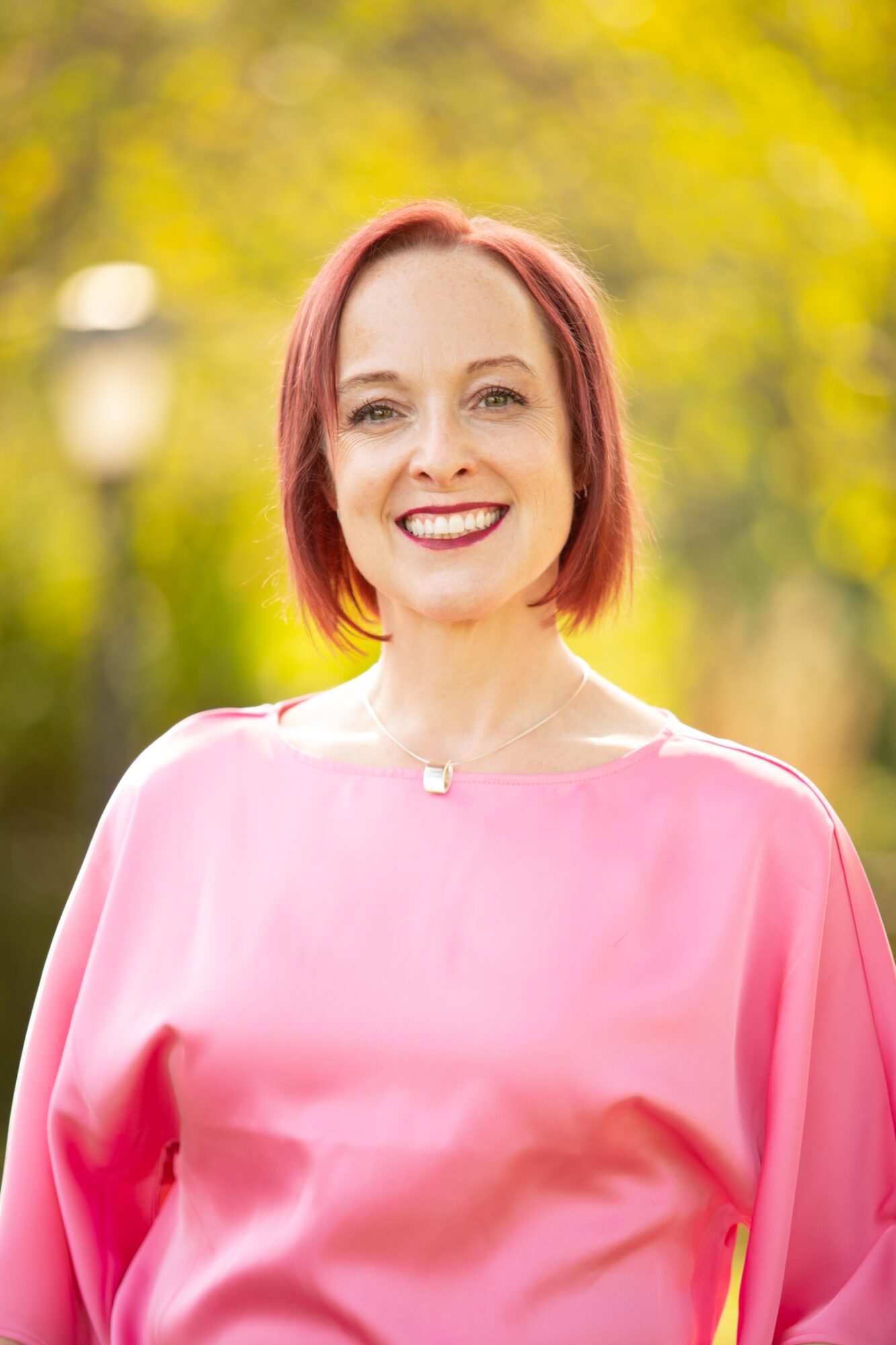 Smiling woman with short red hair outdoors, wearing a pink top, with yellow and green blurred background.