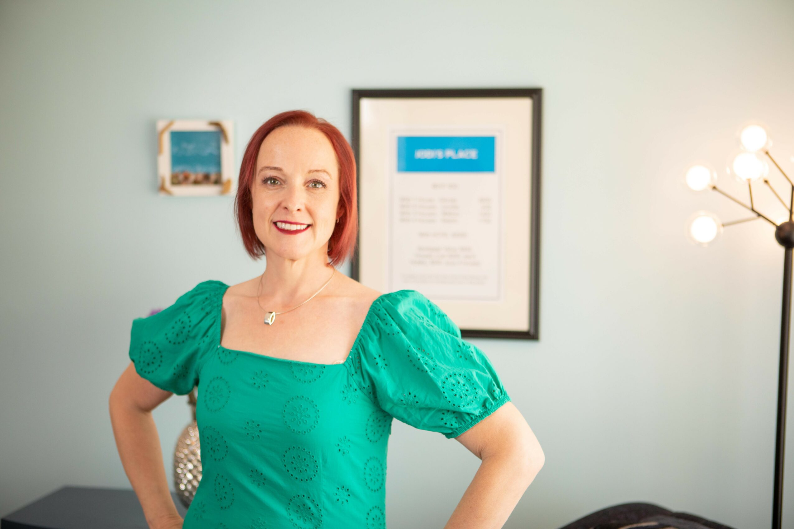 Woman with red hair smiling, wearing a green dress with puffed sleeves, standing in a room with framed artwork and a lamp