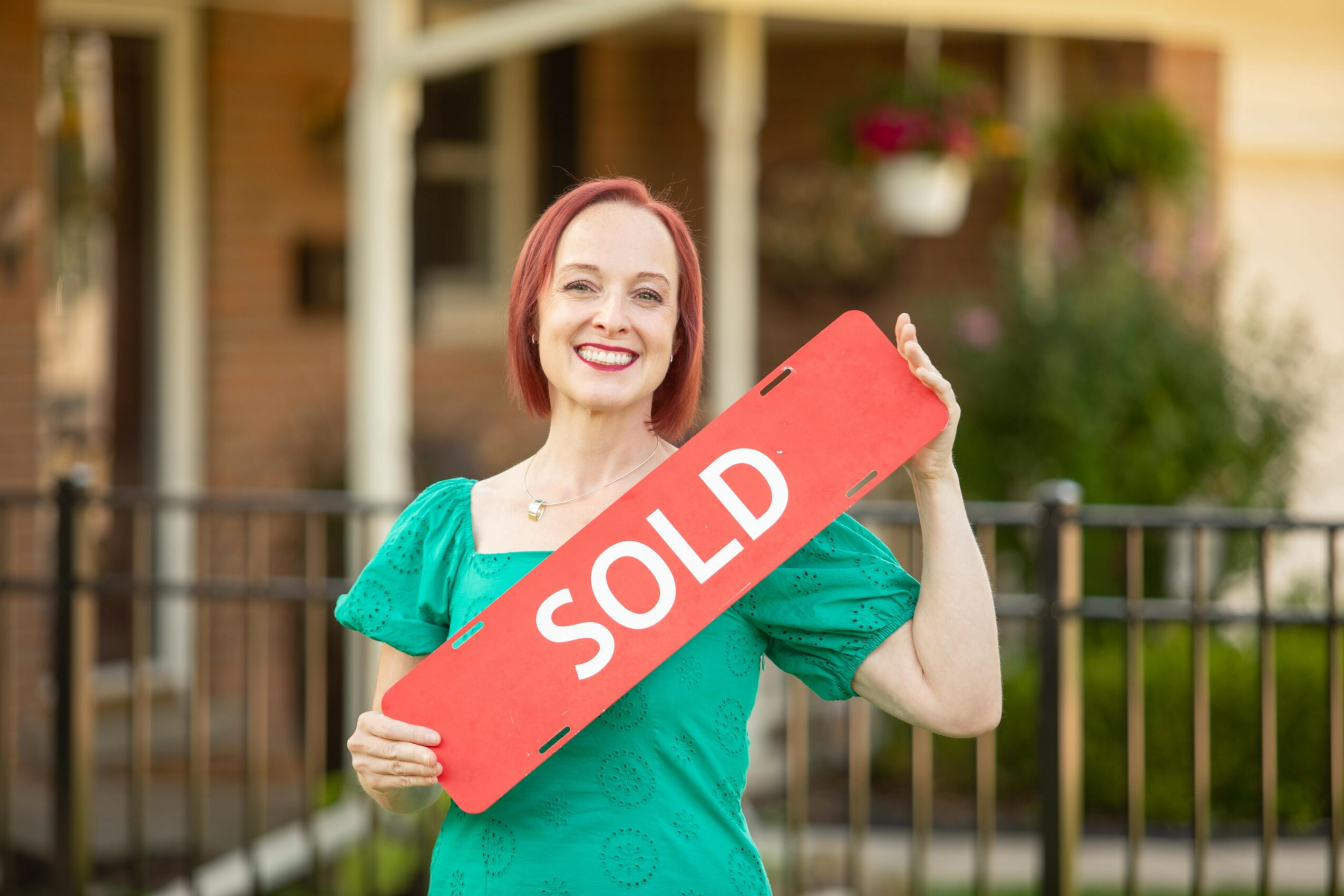 Woman holding a red 'SOLD' sign smiling outdoors in front of a house with a fence and greenery.