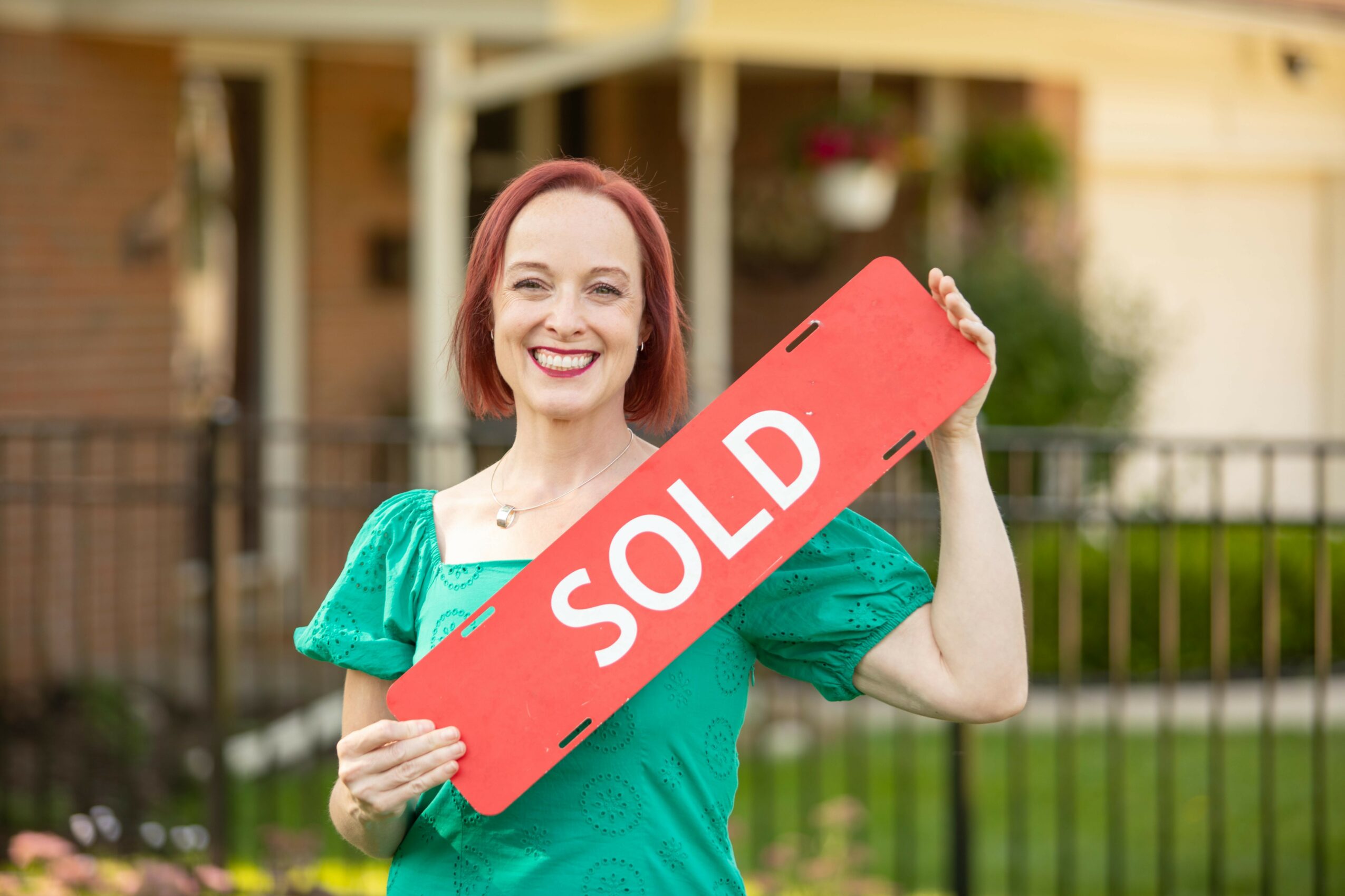 Woman holding a red 'SOLD' sign smiling outdoors in front of a house and fence.