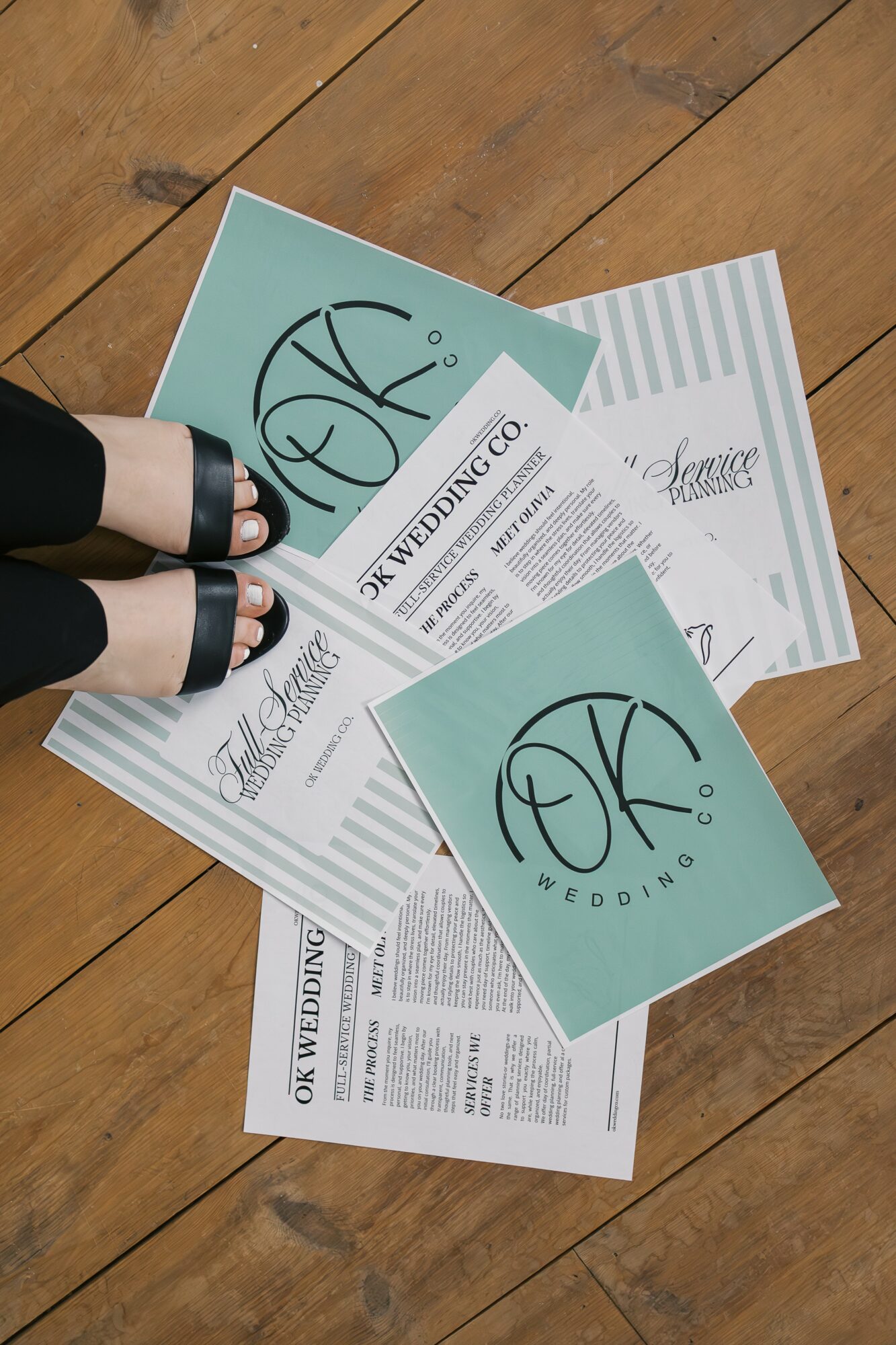 Person's feet in black shoes and black pants stand on scattered wedding brochures and cards on wooden floor.