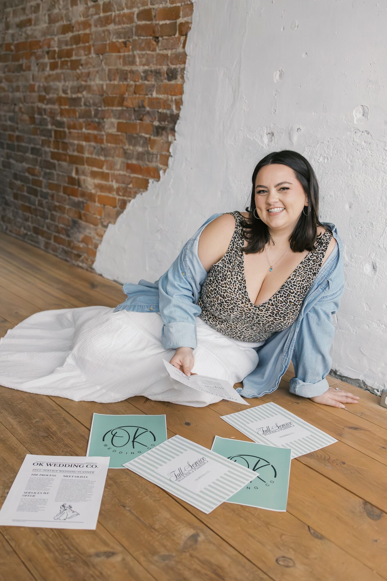 Woman sitting on floor with papers, smiling, against brick and white wall, wearing a patterned top and denim jacket.