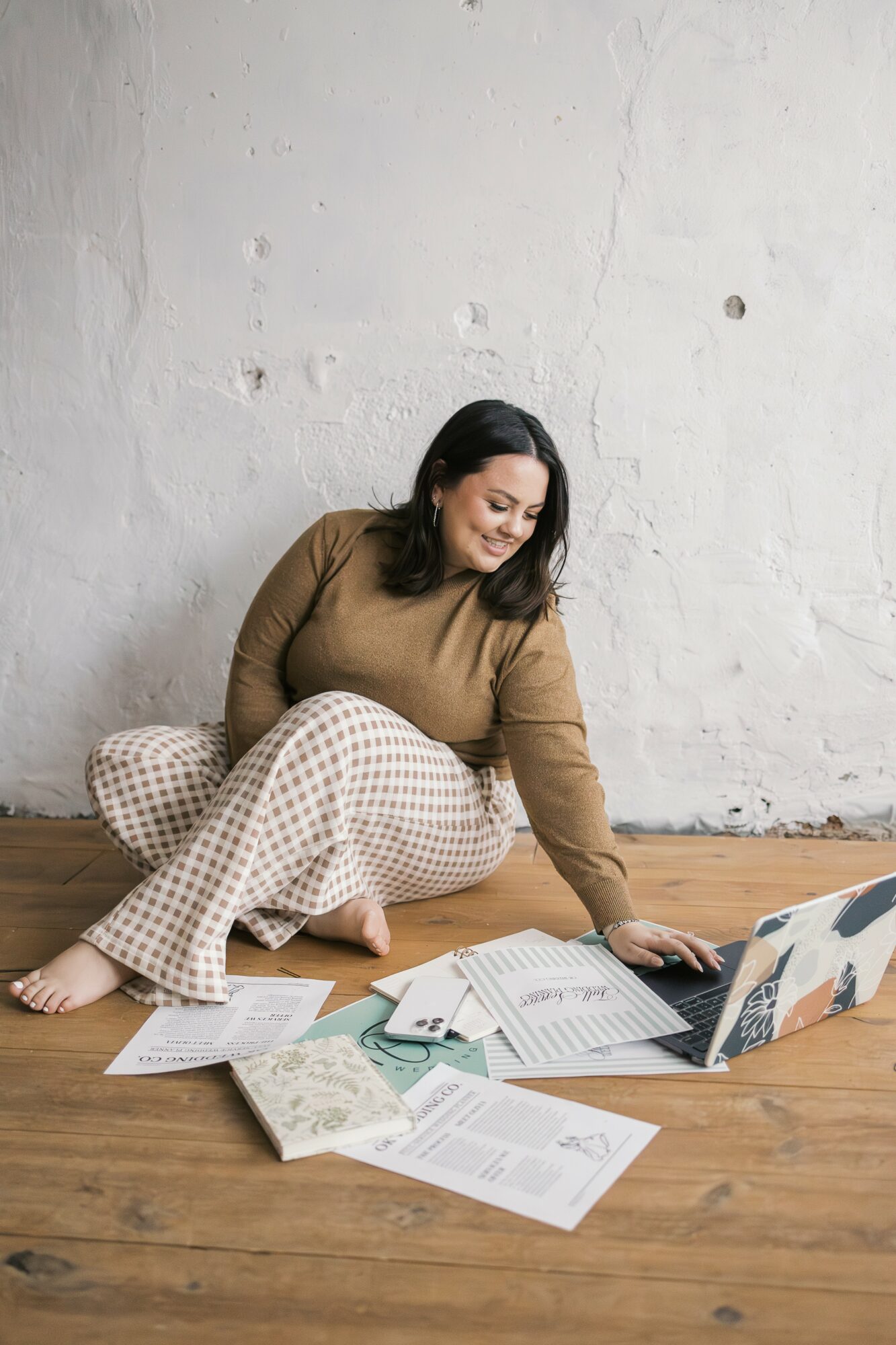 Woman sitting on wooden floor surrounded by papers and a laptop, leaning forward and smiling, against a plain white wall.