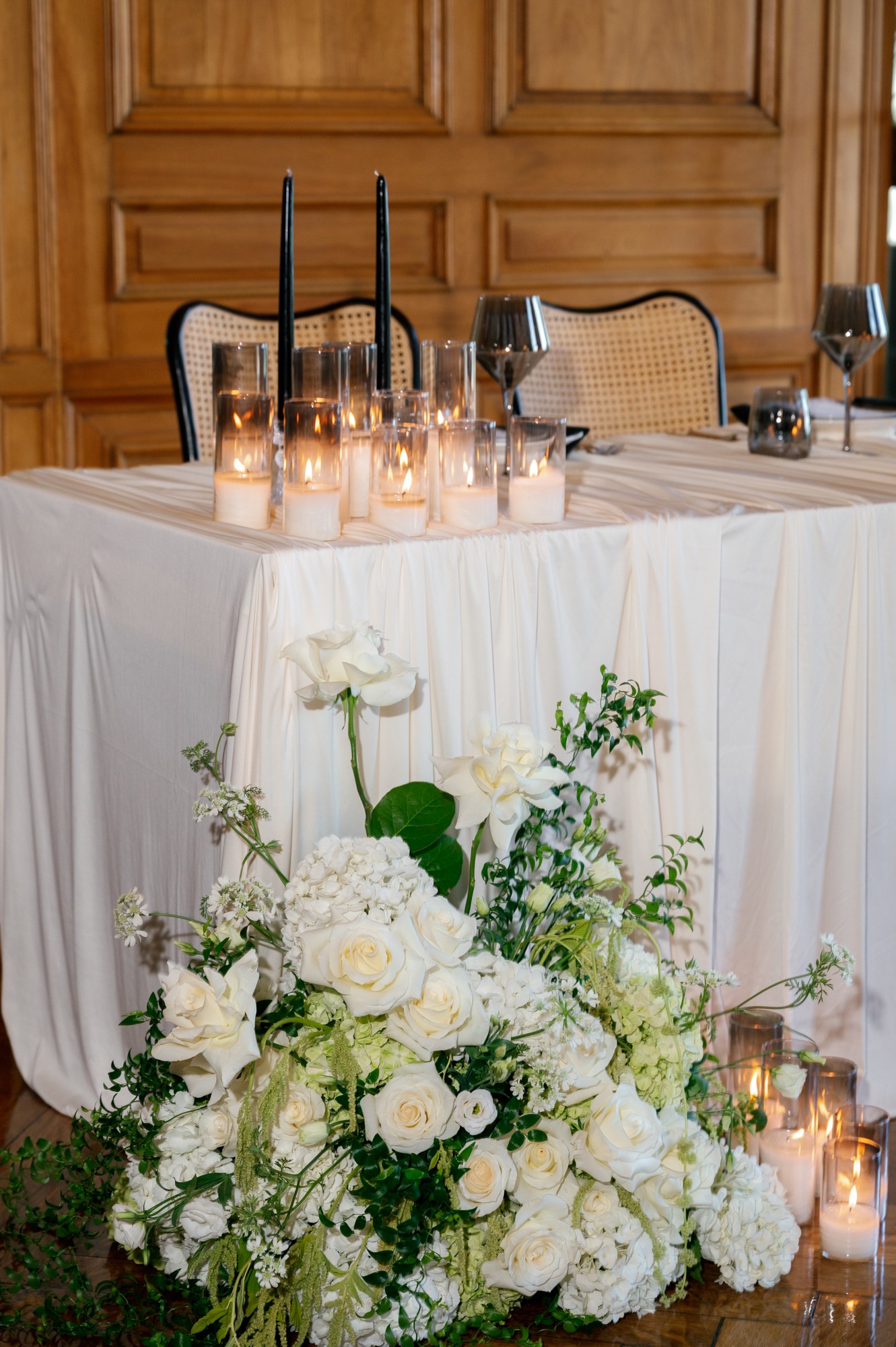 Table with candles and floral arrangement of white flowers and greenery, set in a room with wooden paneling.