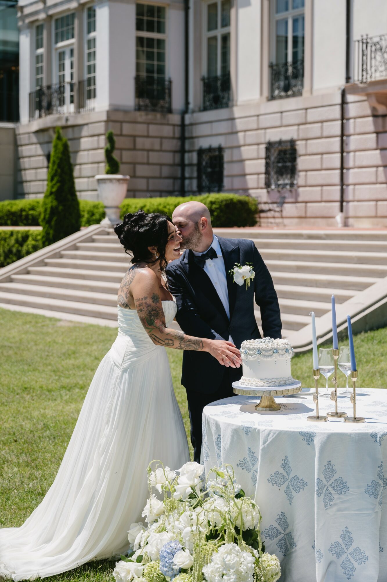 Bride and groom kiss outdoors near a wedding cake, with a grand building and garden in background.