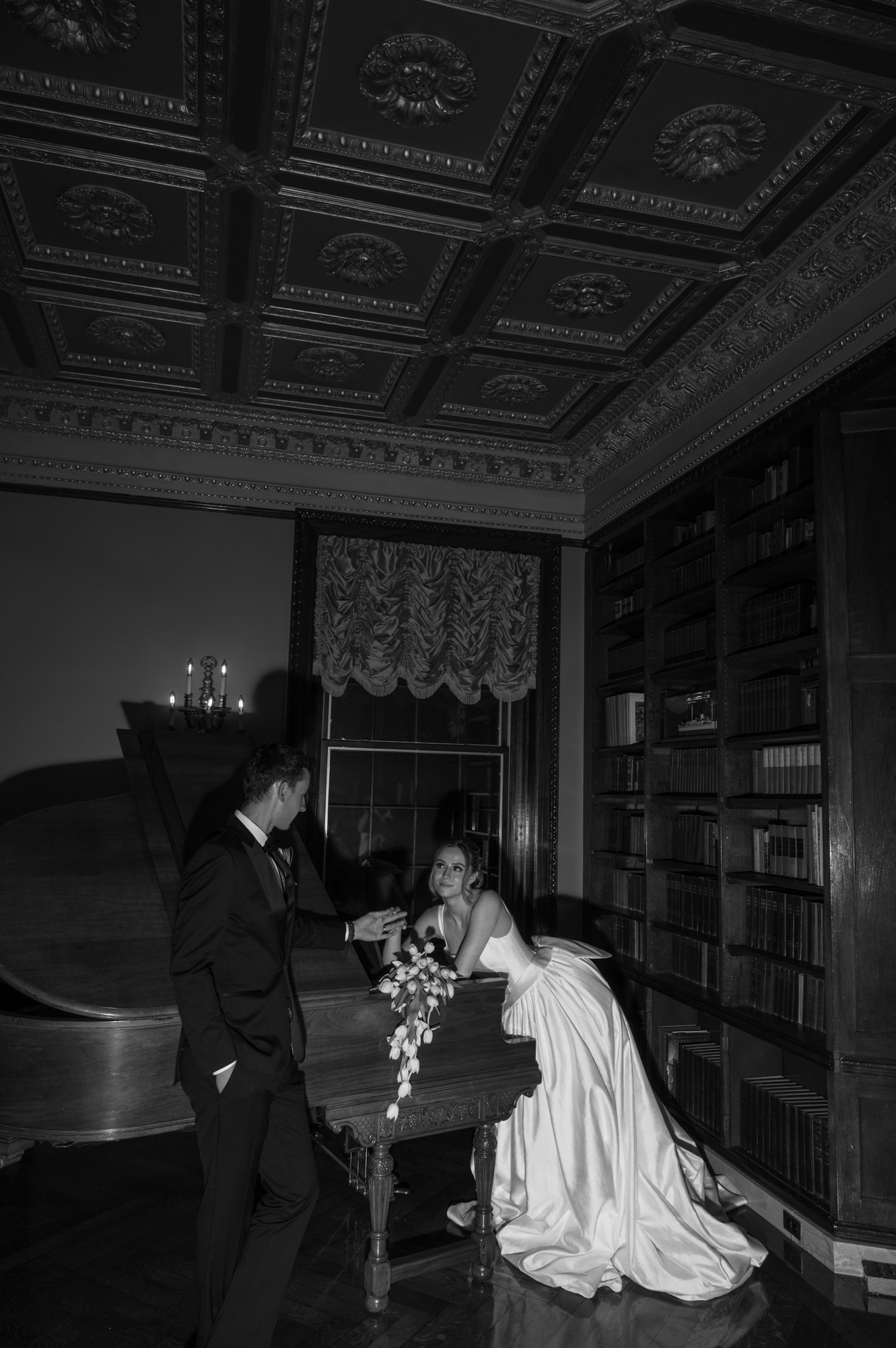 Two women in formal dresses interact in a room with ornate ceiling, bookshelf, and window with lace curtain.
