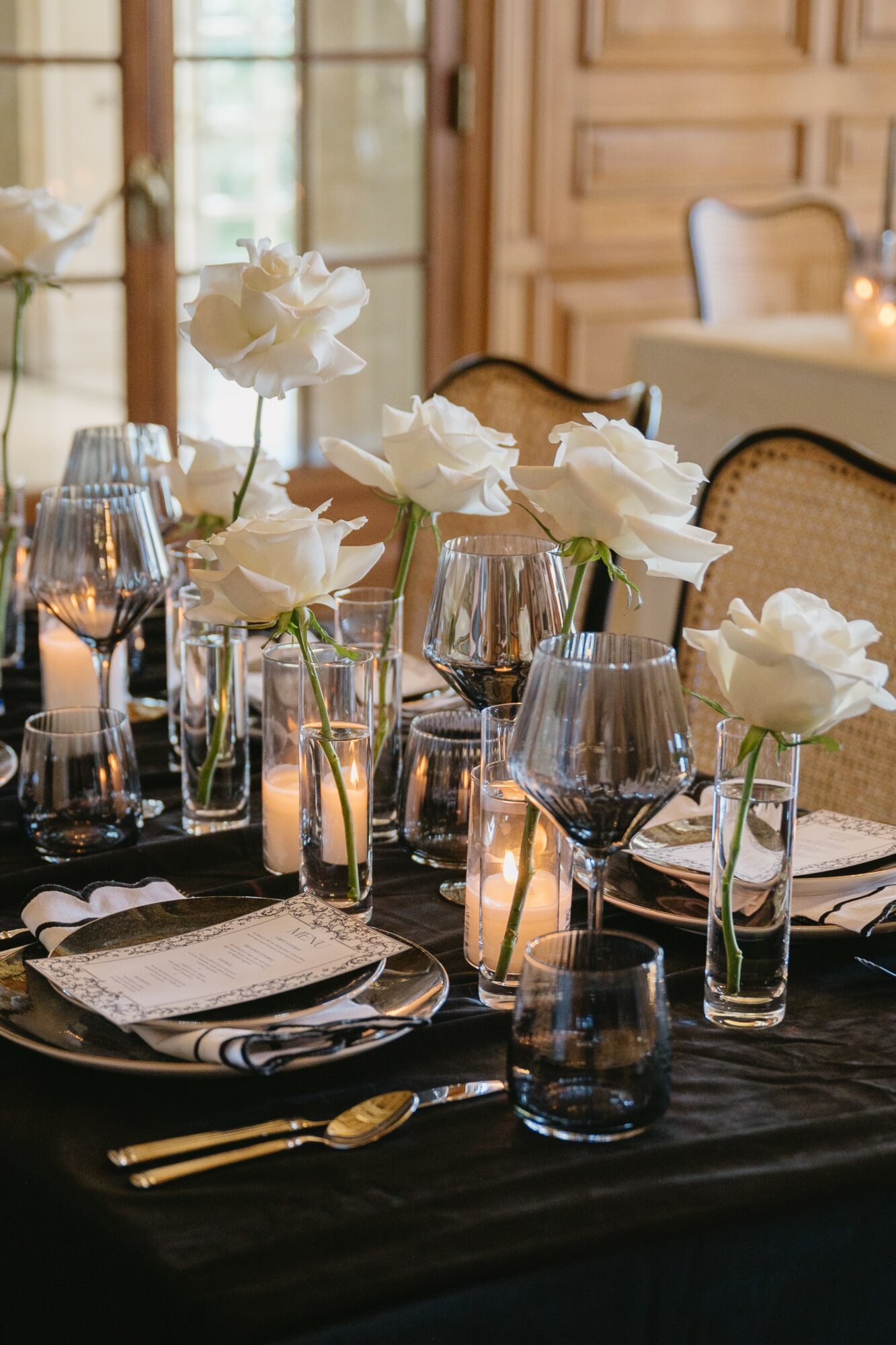 Elegant table setting with white flowers in vases, wine glasses, candles, and black tablecloth in a wooden room.