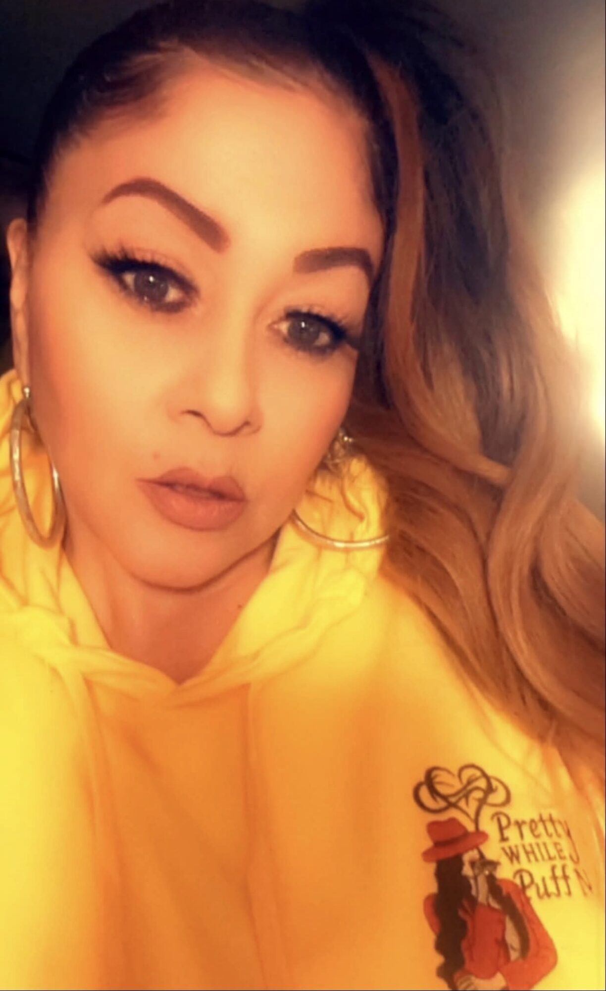 Woman with makeup and large hoop earrings wearing a yellow hoodie with a graphic design, looking at camera.