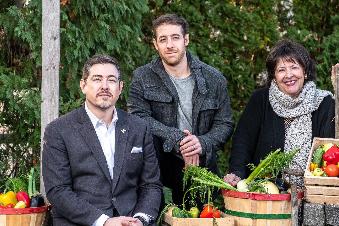 Three people standing outdoors with greenery, smiling, surrounded by baskets of vegetables and fruits.