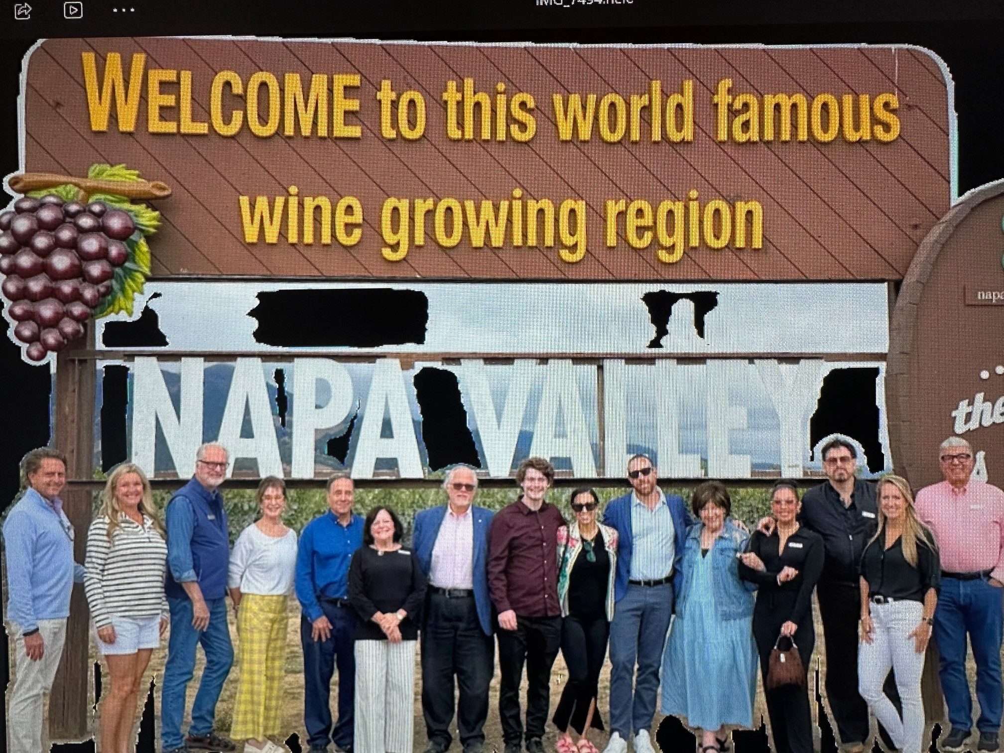 Group of people standing in front of a sign that says 'NAPA VALLEY' and a welcome sign for a wine region.