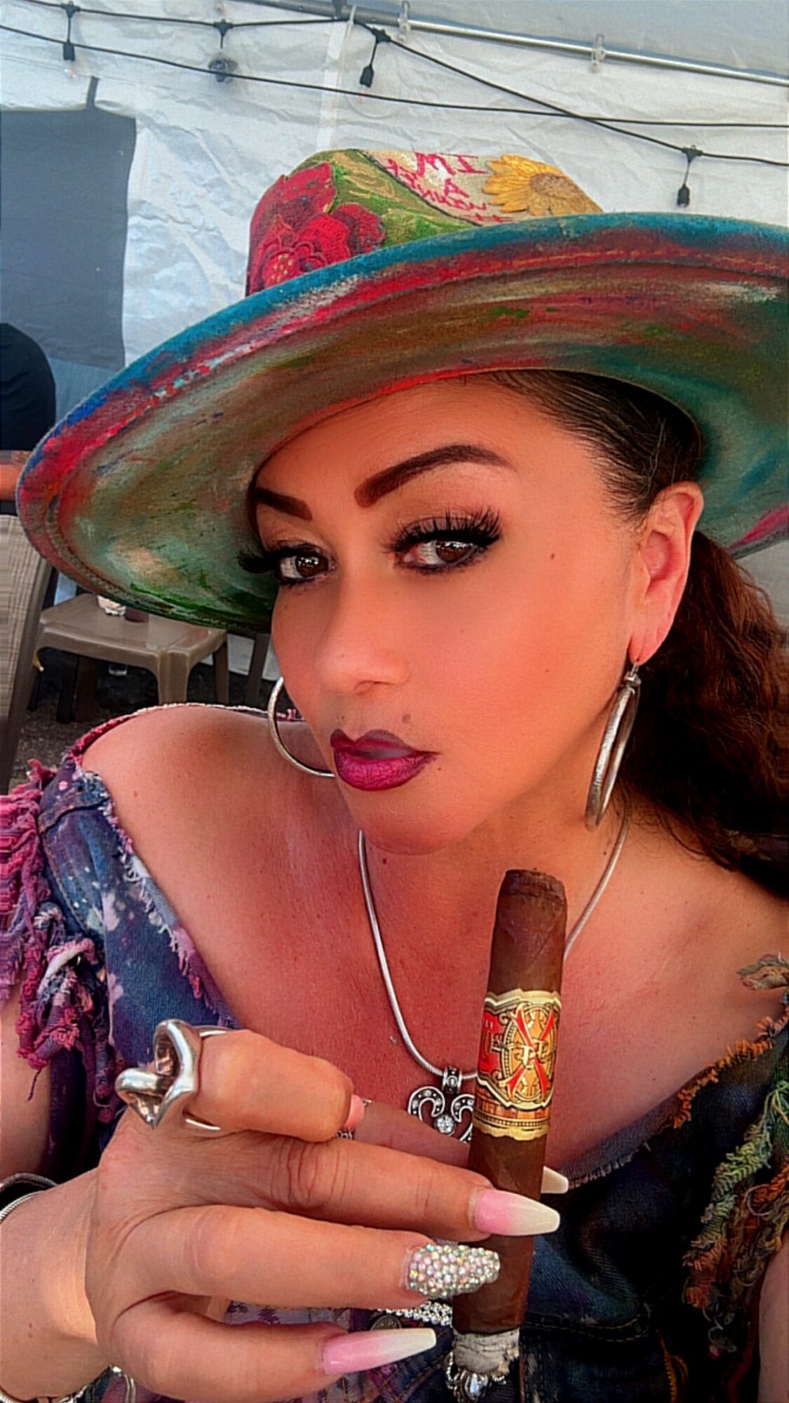 Woman wearing a colorful hat, holding a cigar, with makeup and jewelry, in a festive setting.