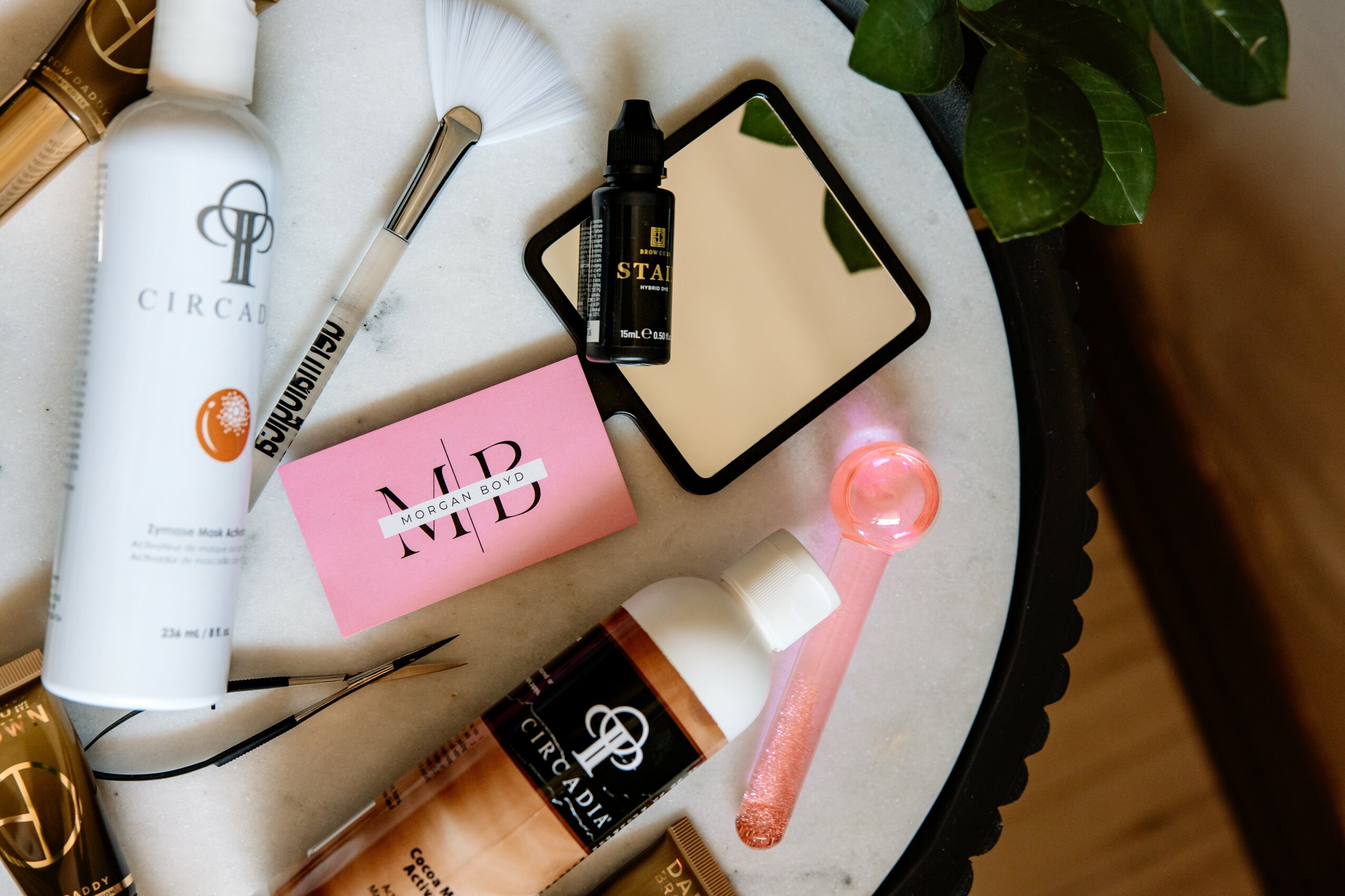 Beauty products, a pink card, a mirror, and a plant on a round table, with a wooden floor nearby.