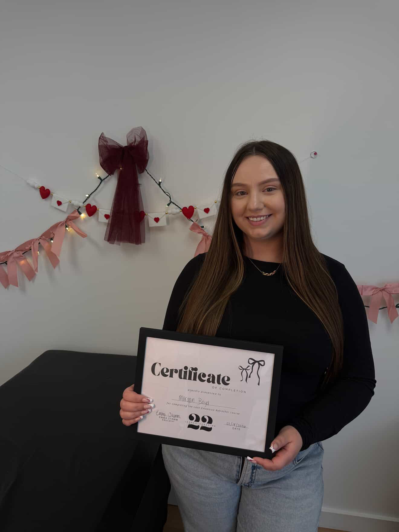 Young woman holding a certificate, smiling, standing in front of a decorated wall with hearts and a bow.