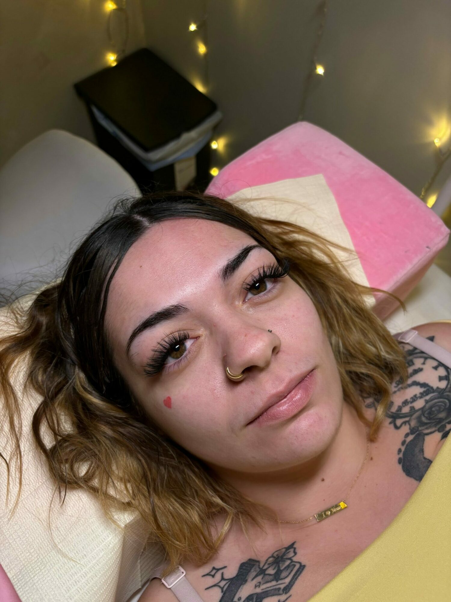 Woman with tattoos and piercings lying on a bed, looking at the camera, with fairy lights in the background.