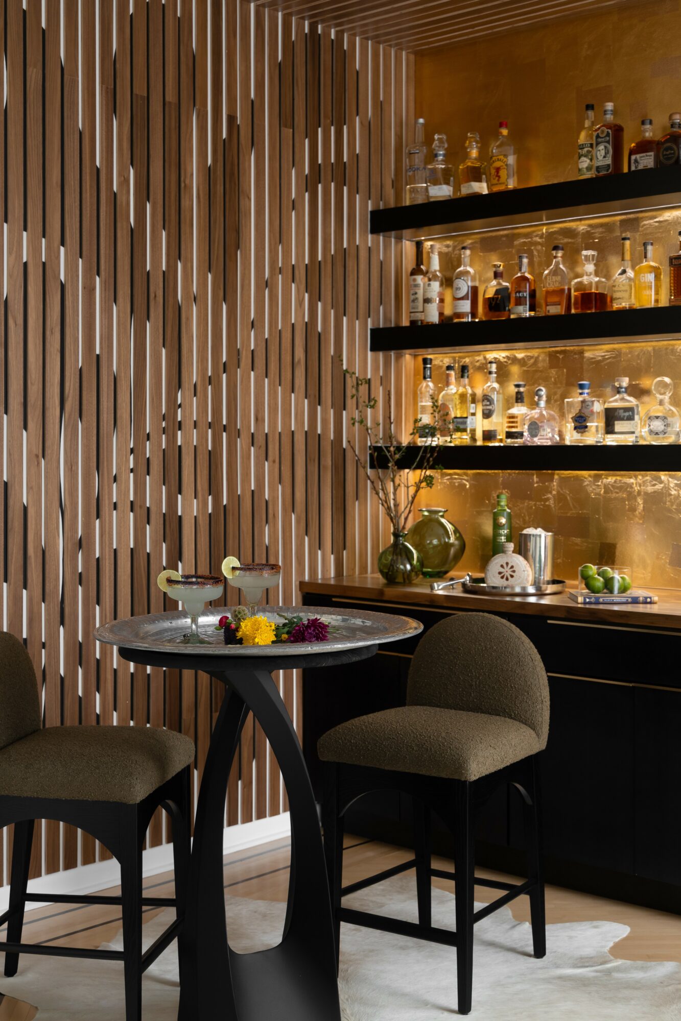 Corner of a bar with a round table, two chairs, and shelves with bottles, against a wooden wall.