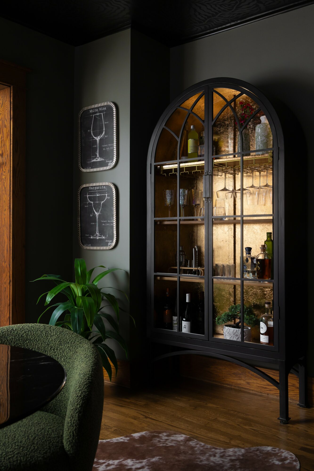 Corner of a room with a black arched glass cabinet, potted plant, and wall art, with wooden floor and dark walls.
