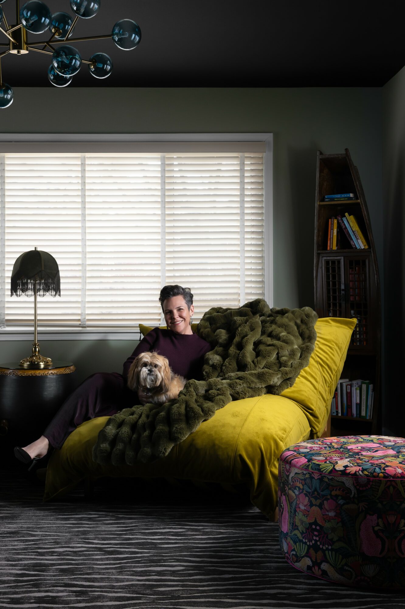 Person sitting on a yellow couch with a dog, in a living room with a window, bookshelf, and patterned ottoman.