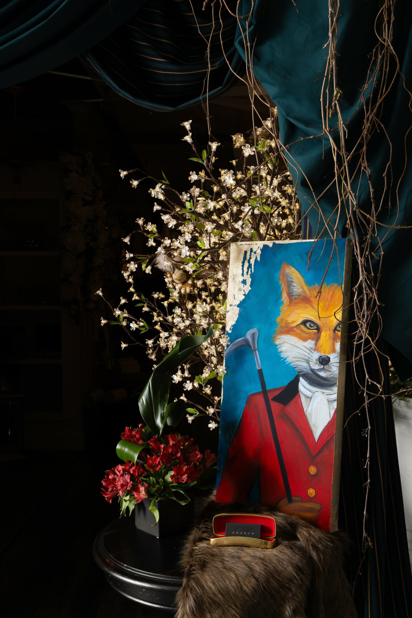 Painting of a fox in a red coat with a blue background, surrounded by flowers and hanging decorations.