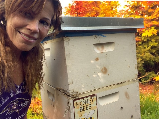 Woman smiling outdoors next to a white beehive with autumn trees in background.