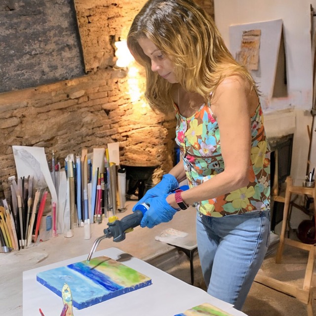 Woman in colorful top and jeans using a heat gun on a small canvas with blue, green, and yellow paint, in a studio.