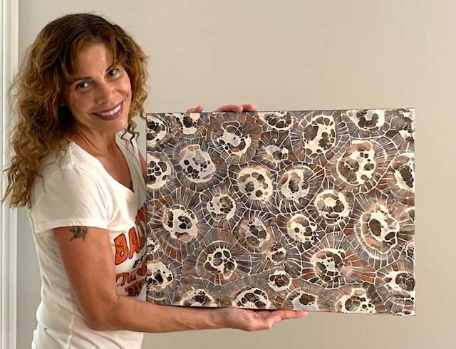 Woman with curly hair holding a large artwork with circular patterns, smiling, standing against a plain wall.