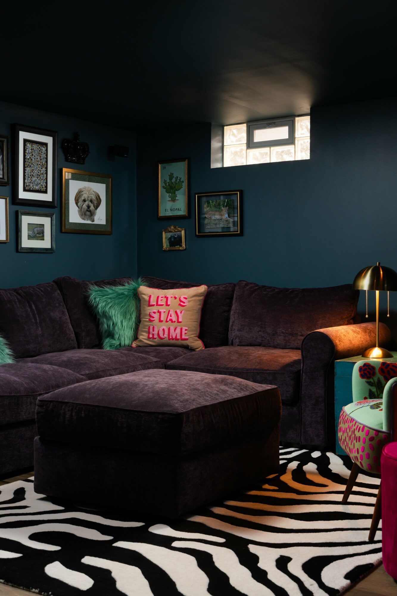 Living room with dark blue walls, framed pictures, a purple sectional sofa, a pink pillow, a zebra-patterned rug, and a small window.