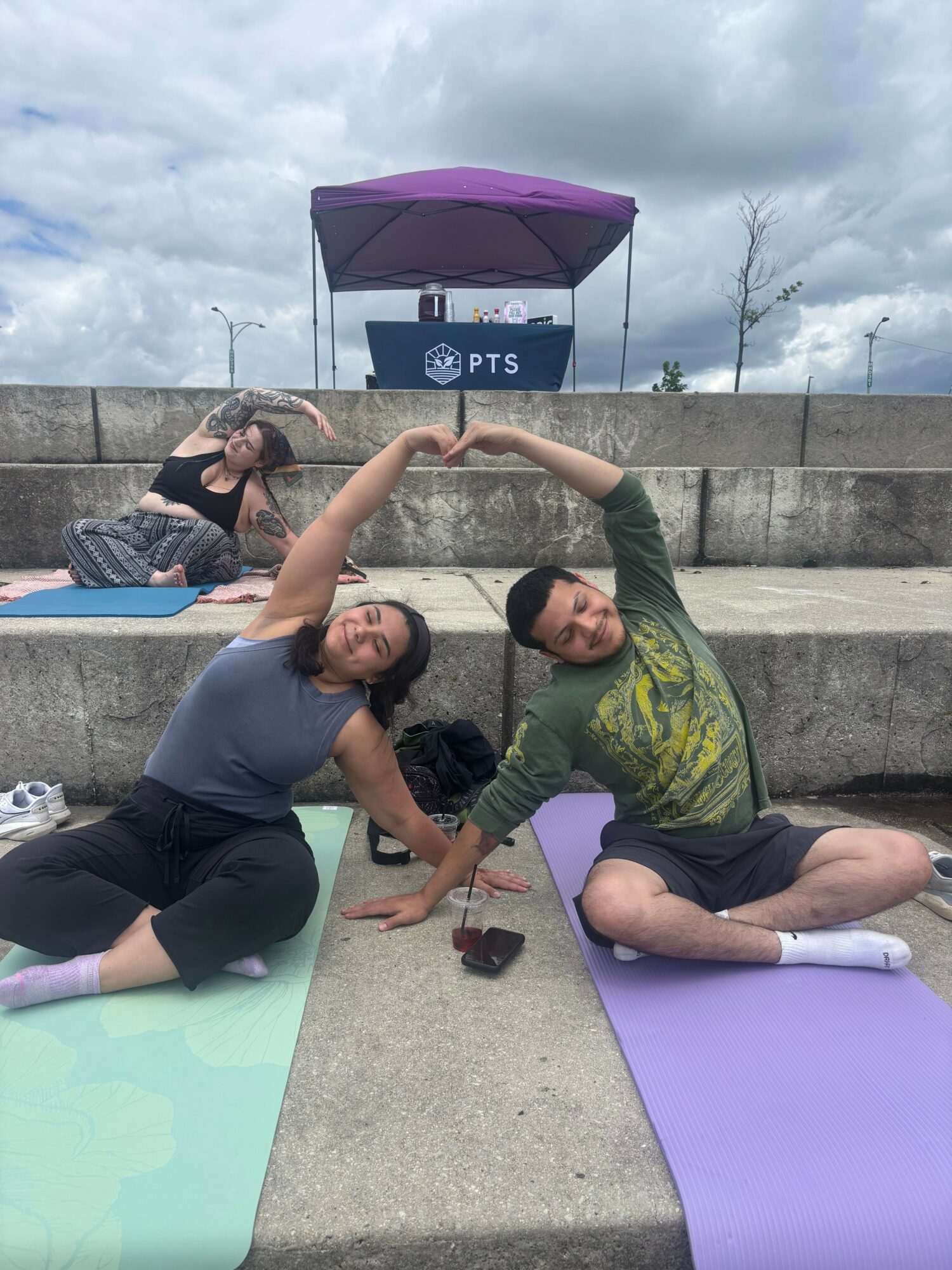 Two women doing yoga outdoors on mats, one holding an umbrella, with a cloudy sky and concrete background.