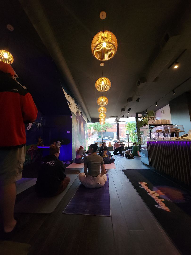 People sitting on mats in a modern indoor space with hanging lights and large windows, some facing away from the camera.