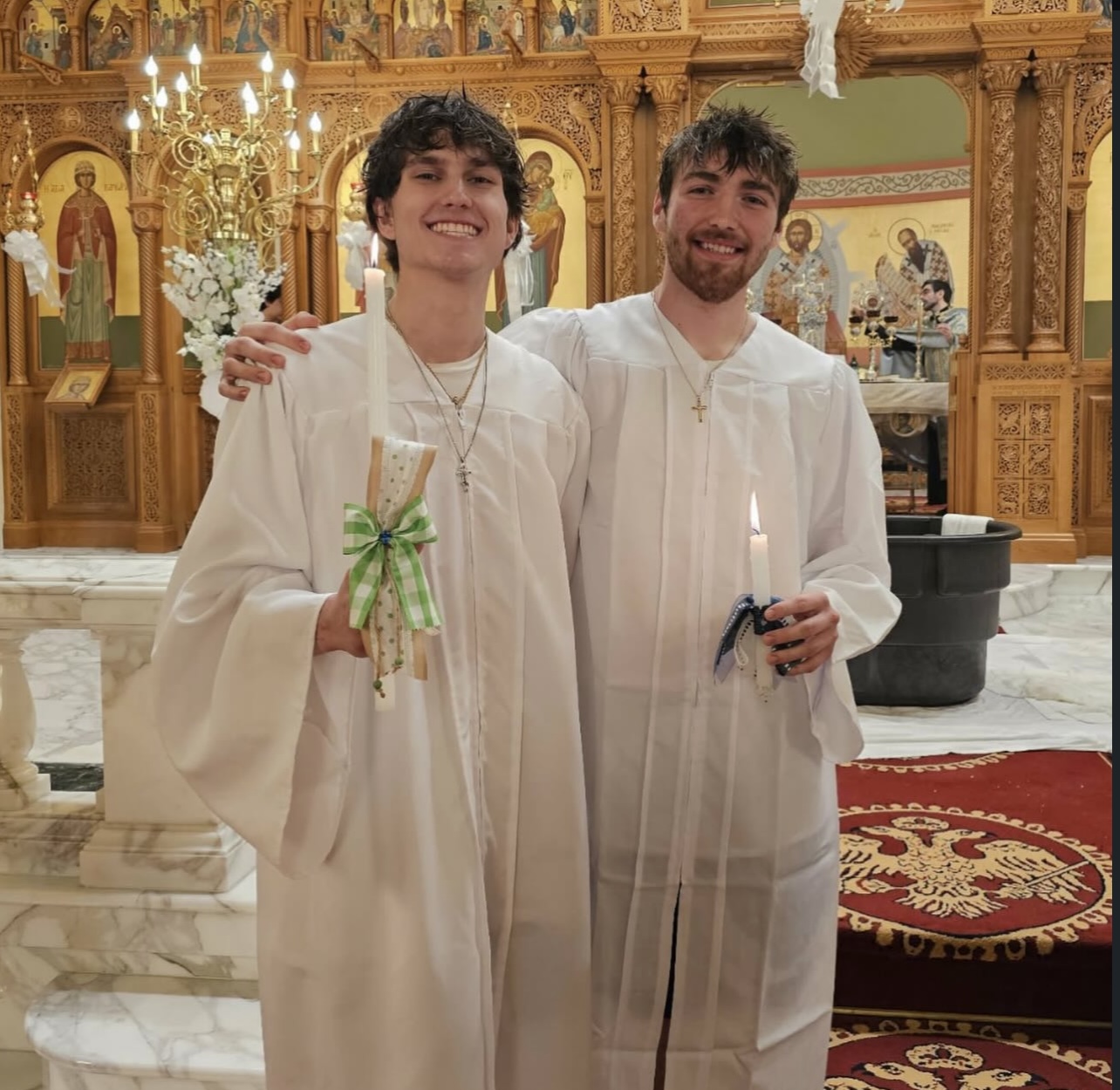 Two young men in white robes standing inside a church, smiling, with ornate gold decorations and chandeliers behind them.