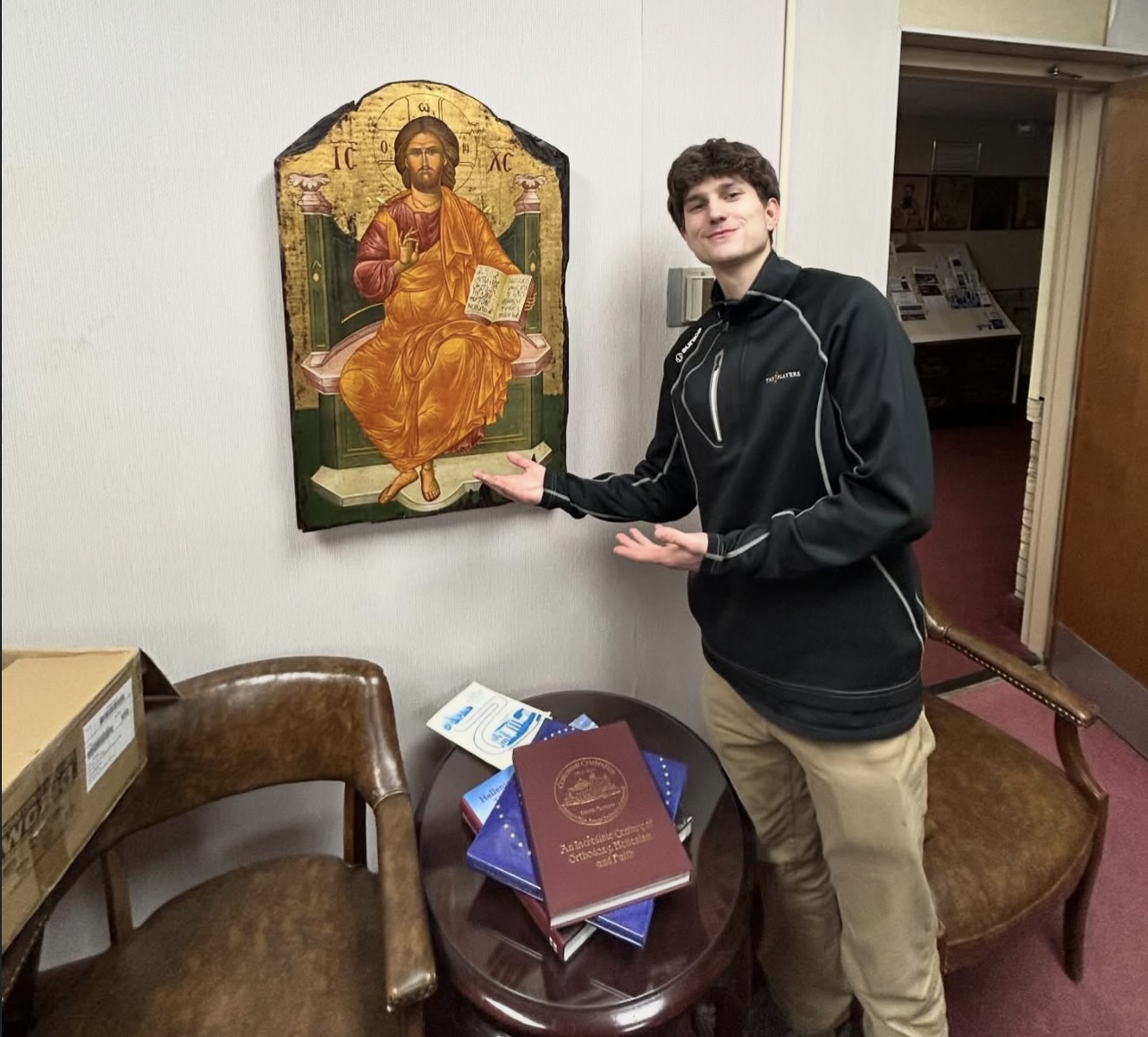 Young man standing next to religious icon, pointing at it, with books on a small table nearby.